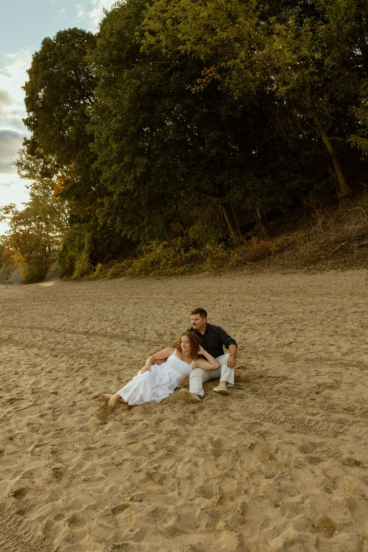 A couple in wedding attire lies together on a sandy beach at sunset with trees in the background.
