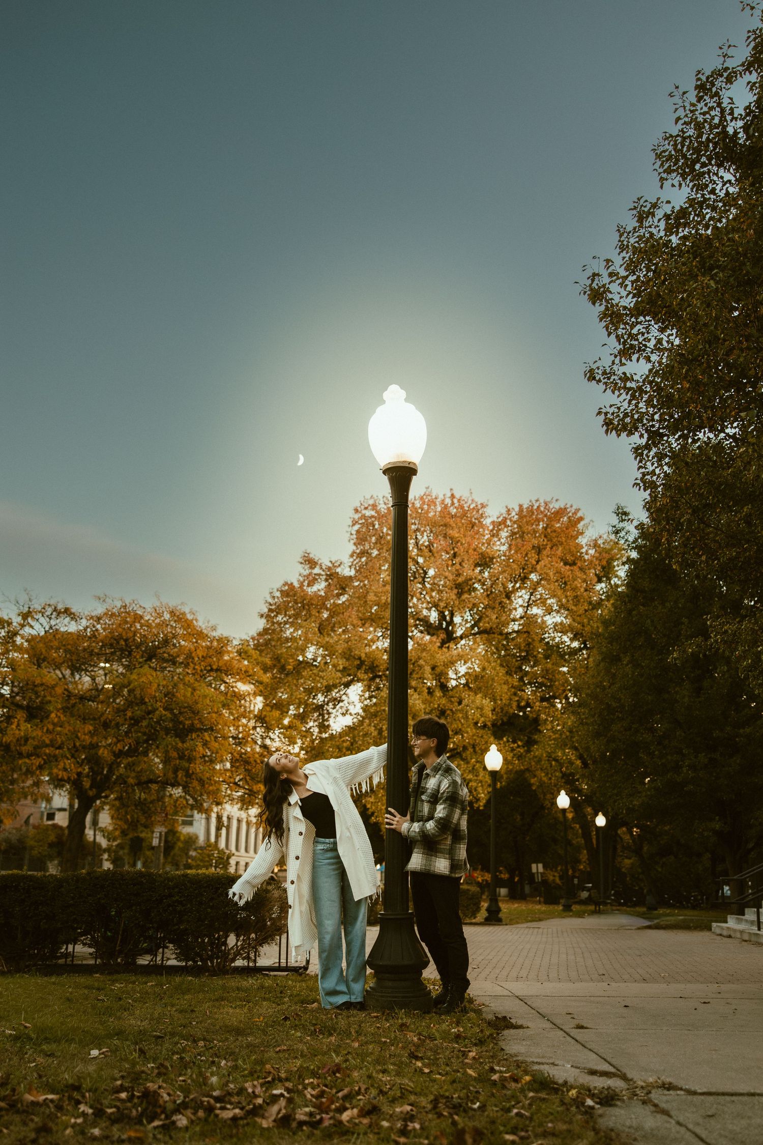 Two people walk along a tree-lined path at sunset with glowing string lights overhead and autumn foliage.