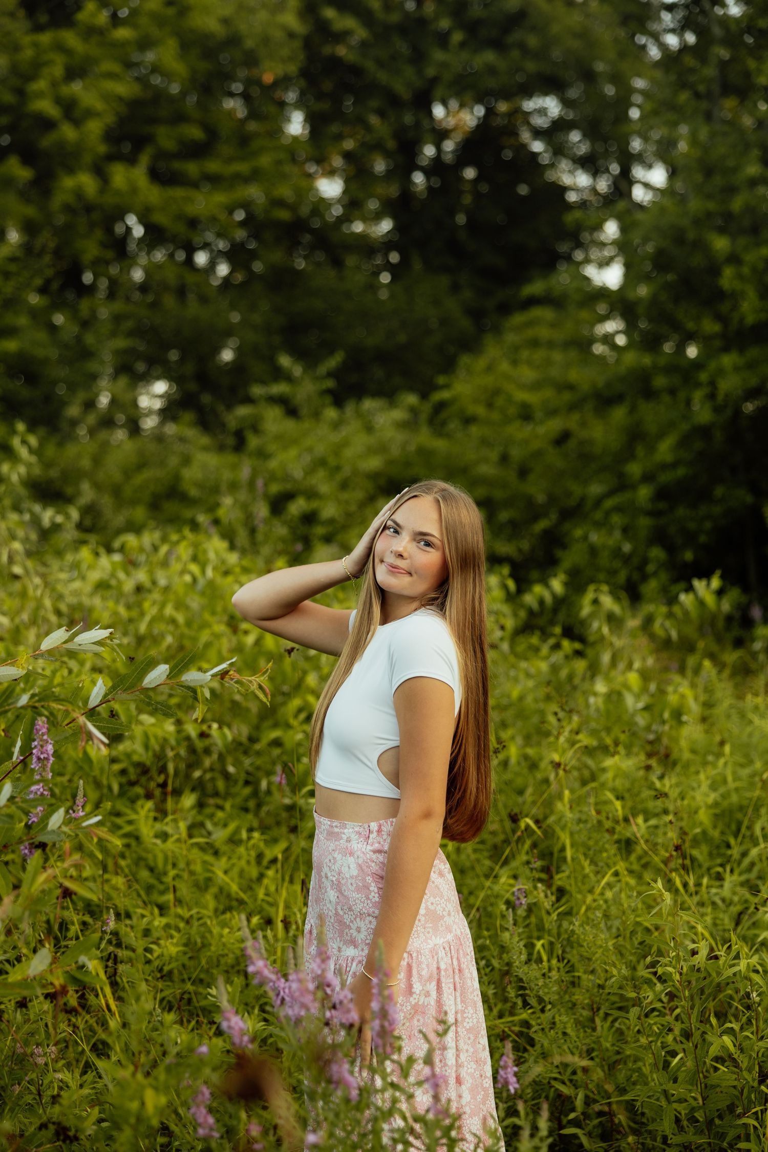 A person in a white crop top and pink floral skirt poses among wildflowers and greenery in a summer garden setting.