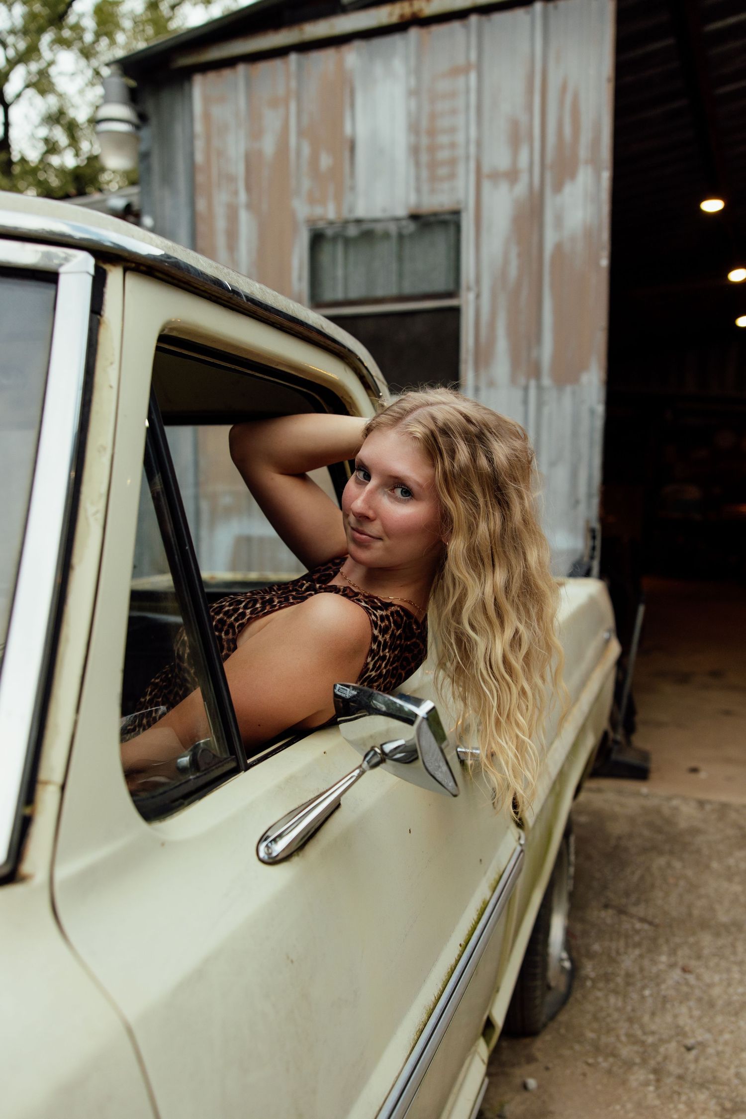 A vintage cream-colored car with a model posing through the window in a rustic outdoor setting.