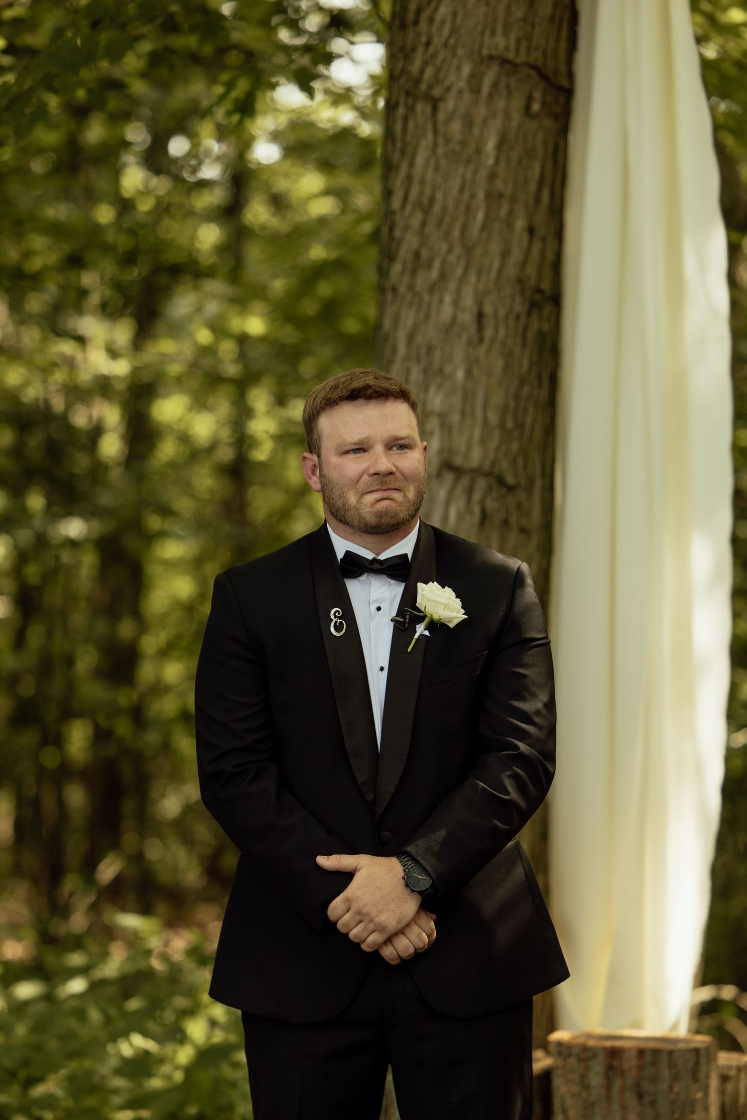 A formal wedding portrait of a groom in a black tuxedo standing in front of a white backdrop outdoors.