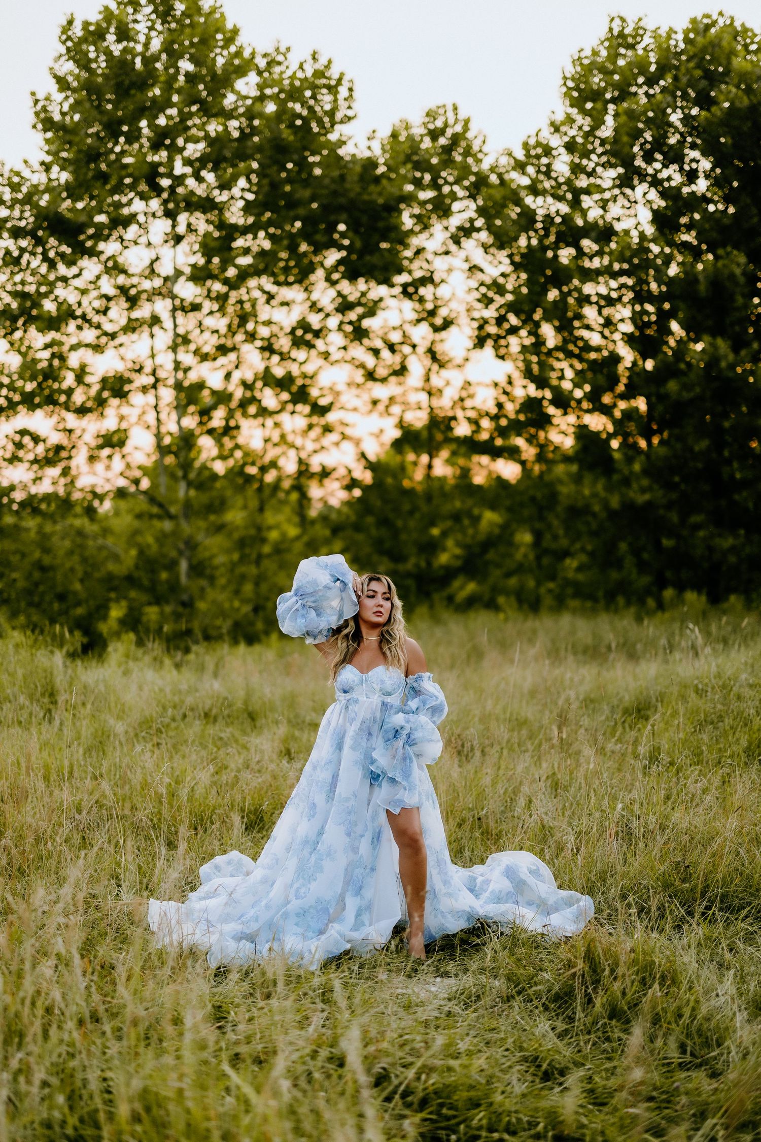 Ethereal portrait in flowing blue gown against sunset in meadow.