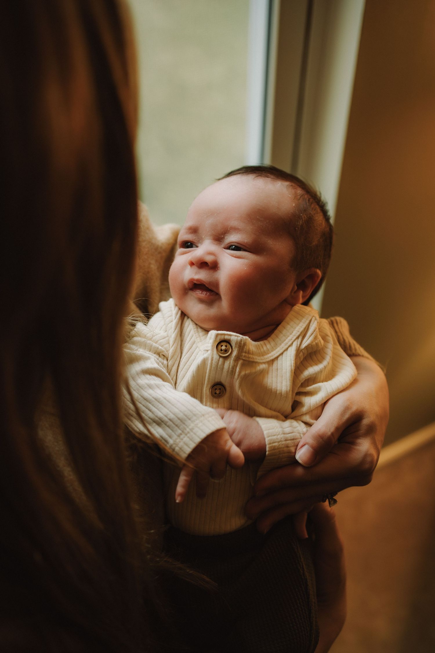 A baby in a cream-colored outfit smiles while being held near a window.