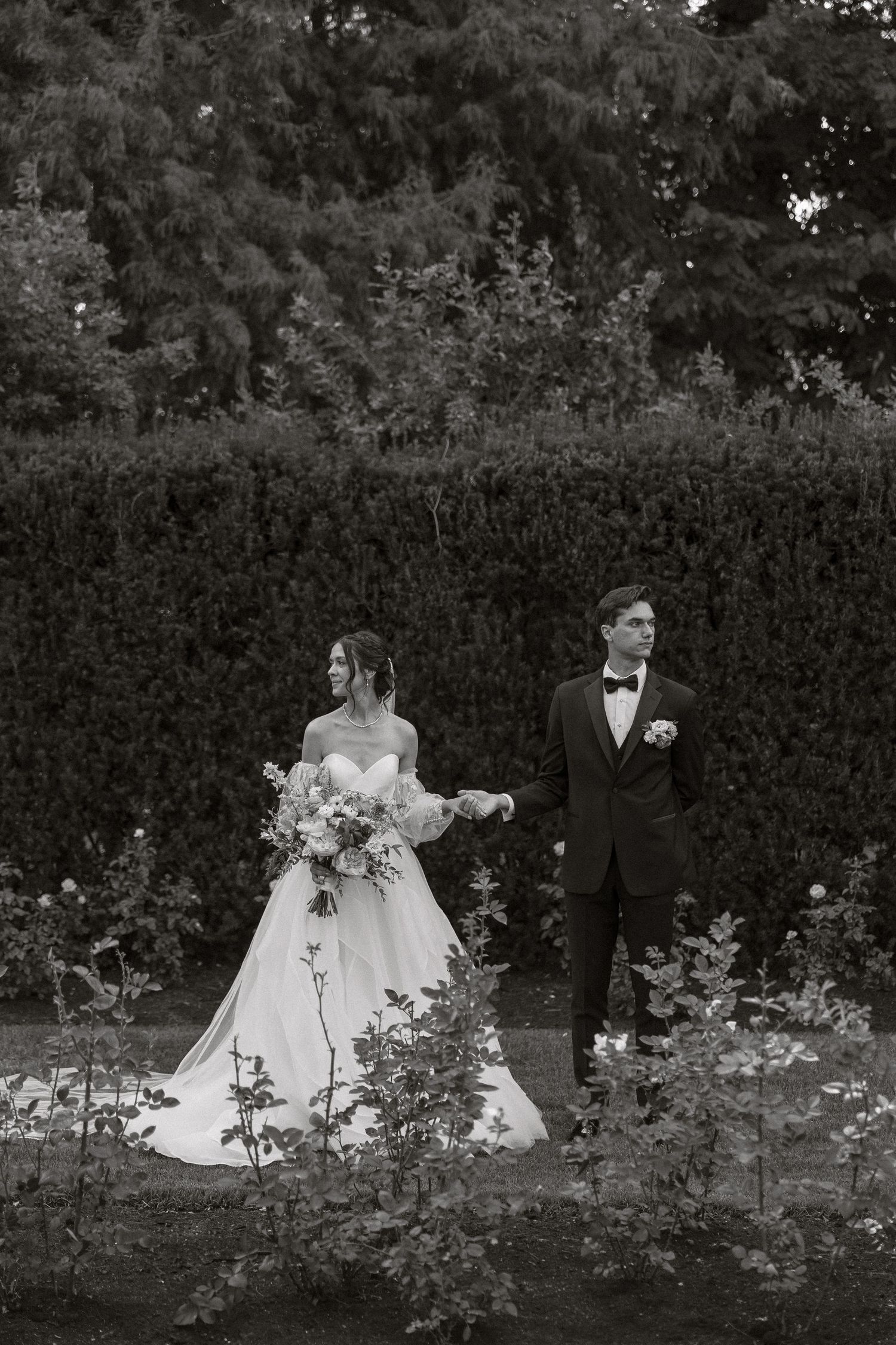A black and white wedding photo of a couple standing in a garden with lush hedges and florals in the background.