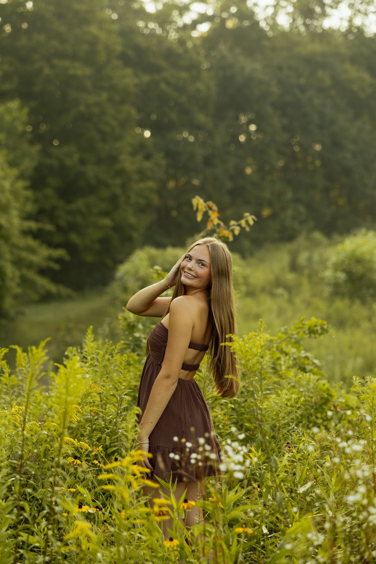 A figure in a brown dress walks through a meadow of yellow wildflowers and tall grass on a summer evening.
