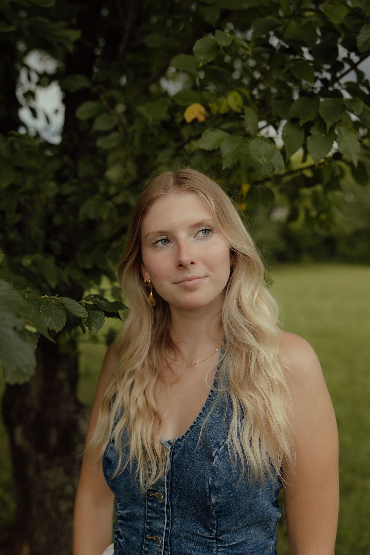 A person in a denim top poses beneath leafy green trees in a natural outdoor setting.