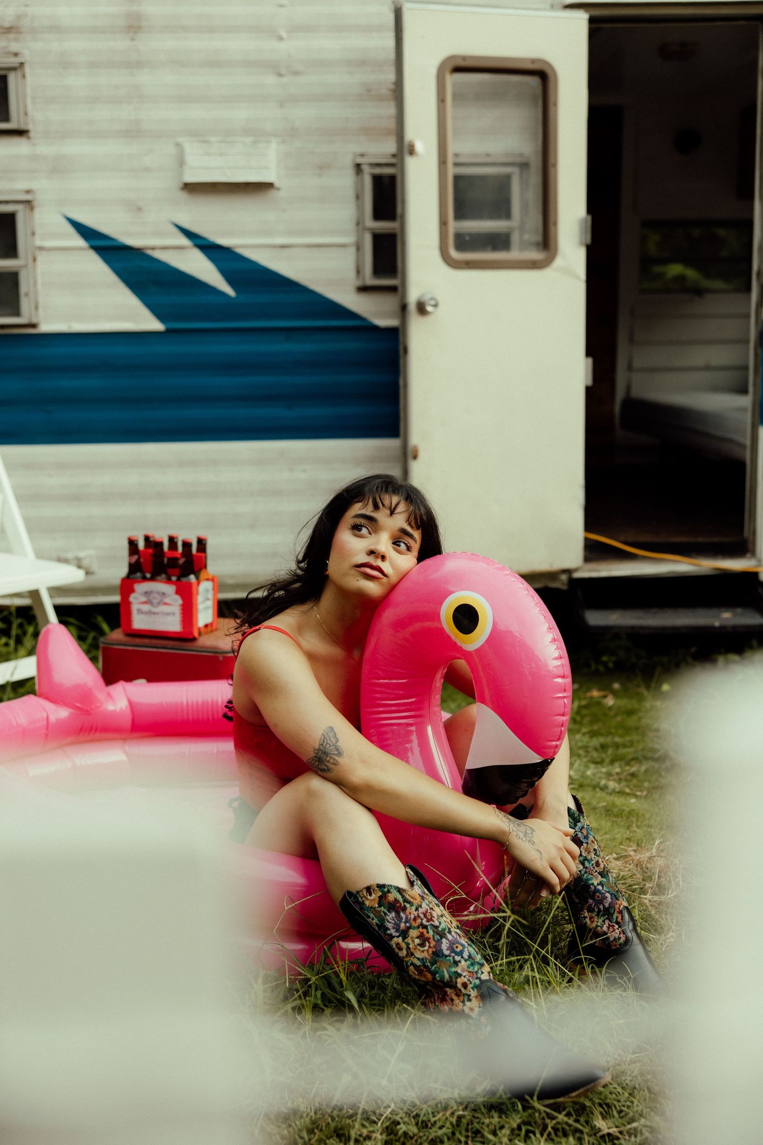 A pink flamingo pool float sits beside a retro white and blue camper trailer in a backyard setting.