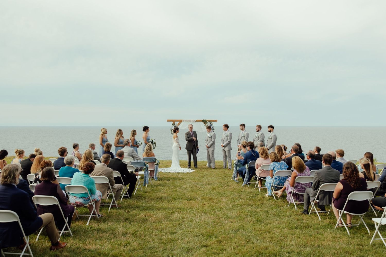 A panoramic view of wedding guests seated in white chairs on a grassy hilltop overlooking the ocean.