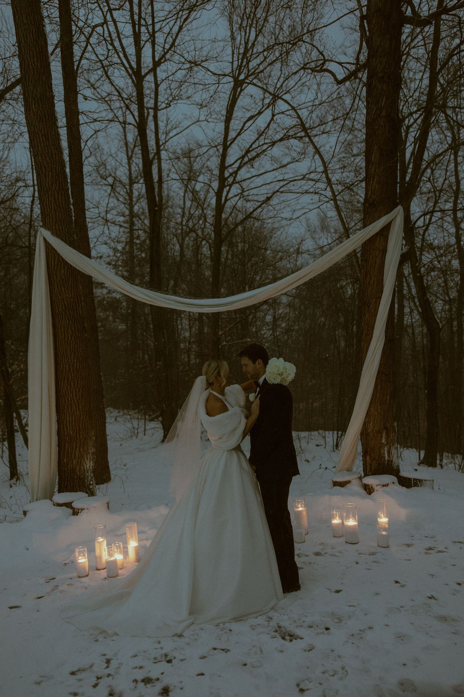 A snowy winter wedding scene with white fabric draping between trees and glowing candles on the ground.