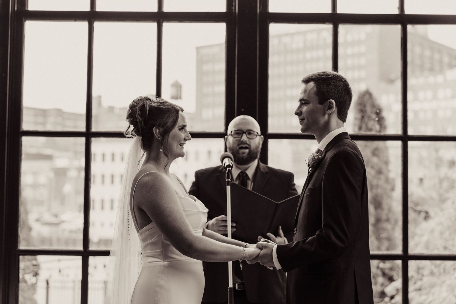 Elegant wedding couple dancing in front of large industrial windows with city views.