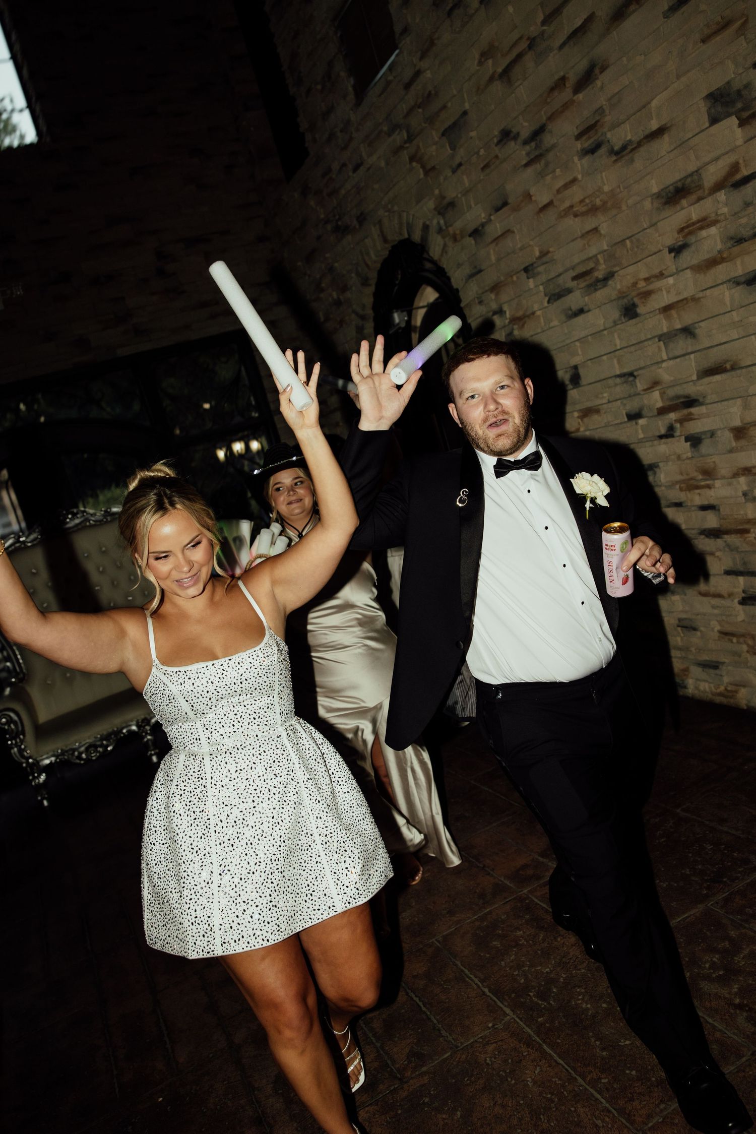 Wedding celebration scene with people dancing and raising their hands in joy against a brick wall backdrop.