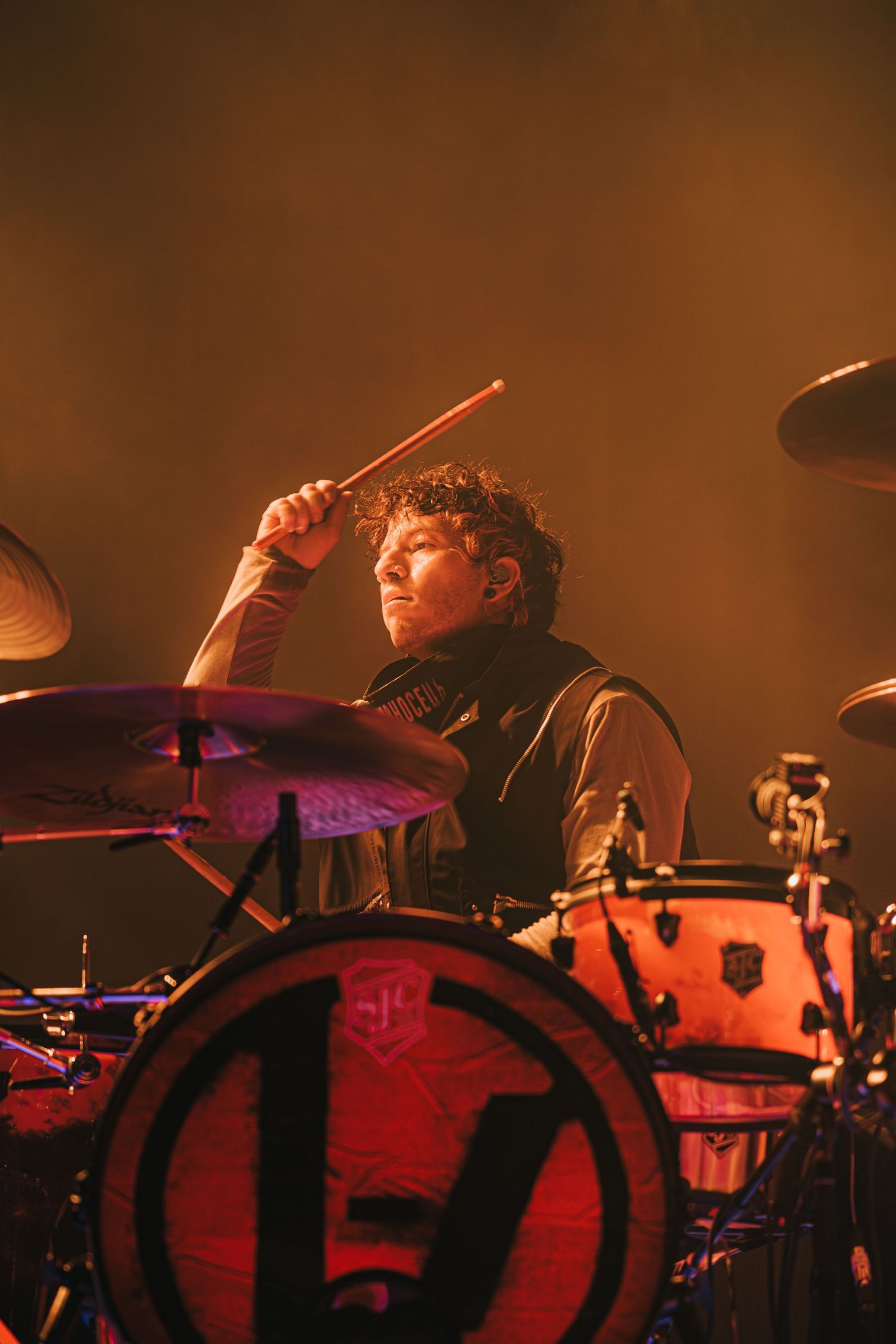 Drummer performs energetically behind drum kit in warm stage lighting during live concert.