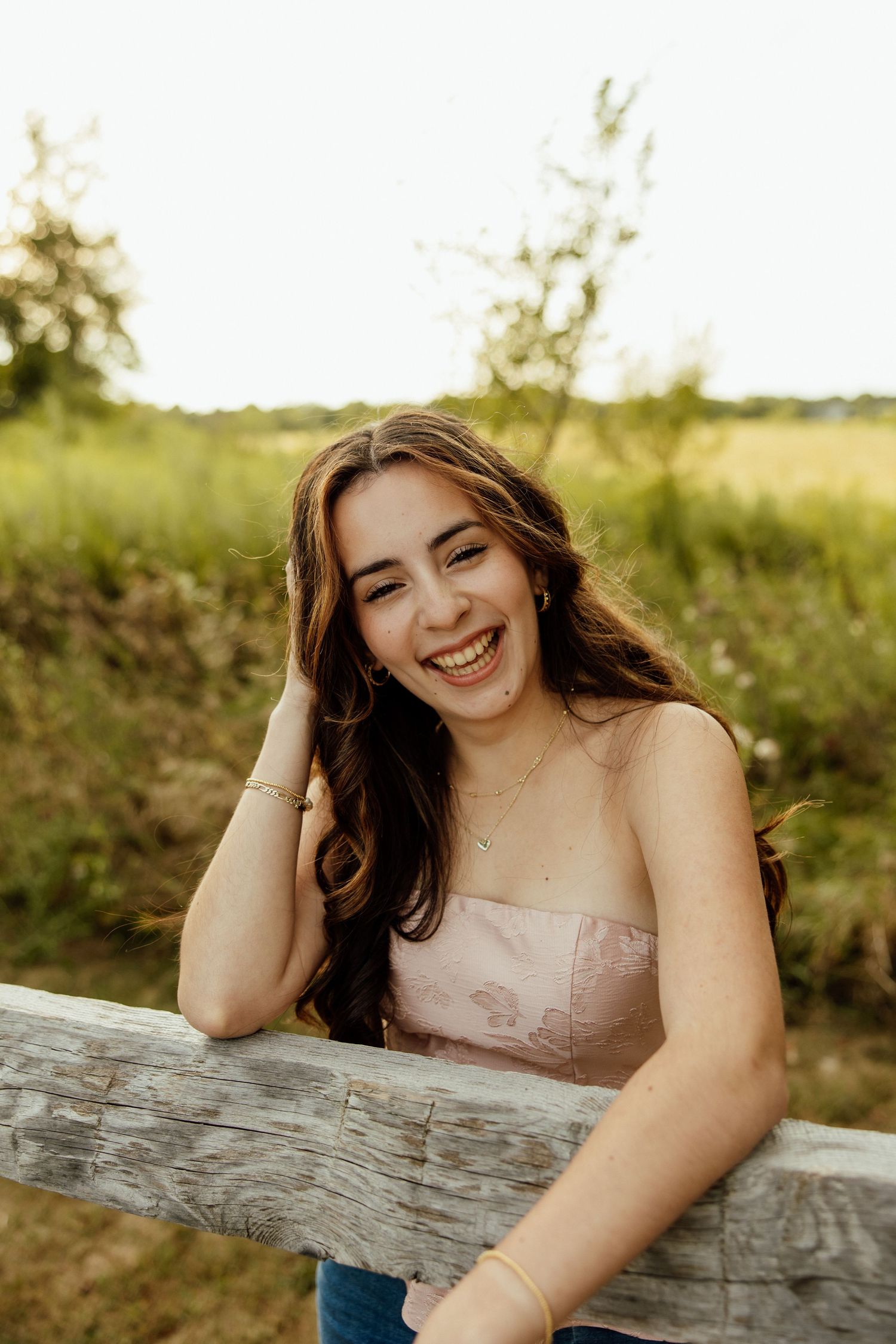A person in a pink dress smiles while leaning on a wooden fence post during a golden hour photoshoot.