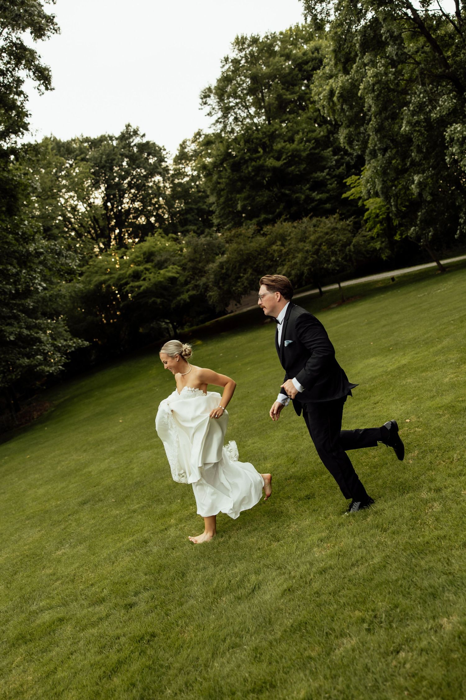 A playful wedding couple runs across a grassy hill during an outdoor photo session.