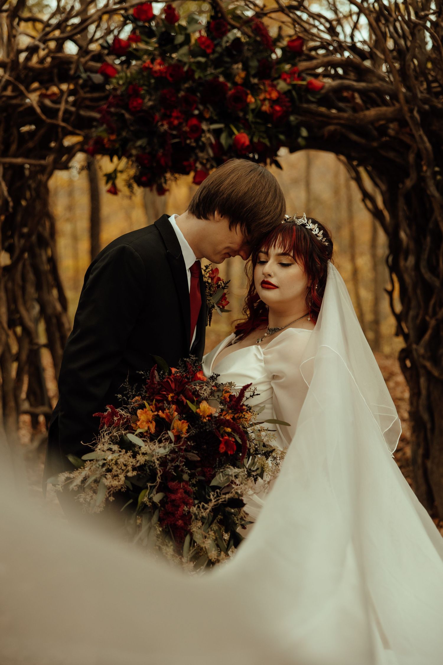 Romantic wedding couple shares intimate moments under rustic wooden arch adorned with deep red flowers and greenery.