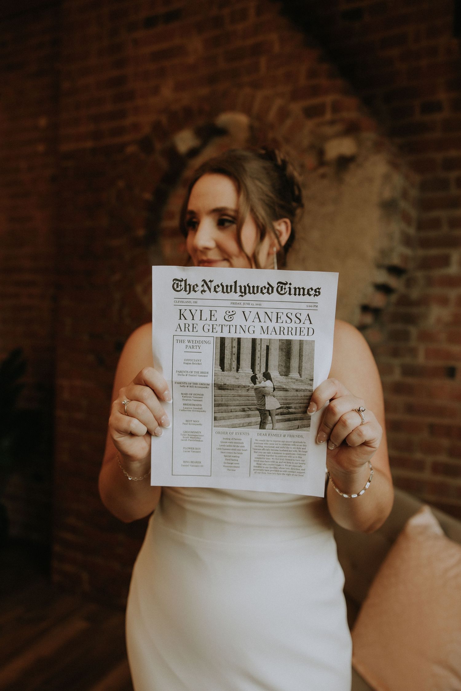 A bride in a white dress holds up a custom newspaper wedding announcement against a brick wall background.