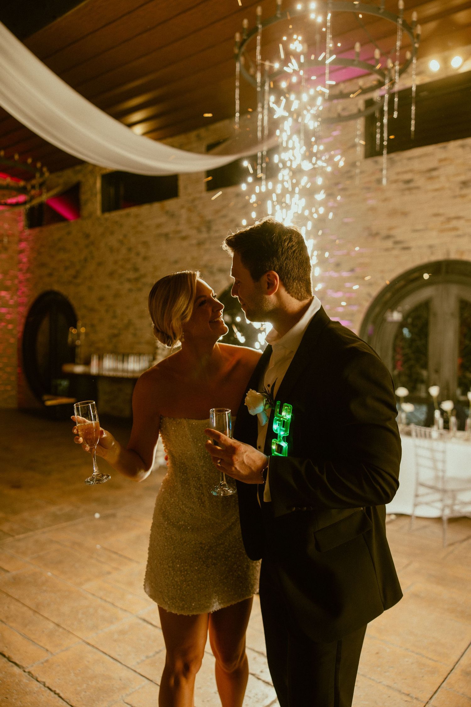 Wedding guests with champagne glasses gather under twinkling lights in an elegant stone-walled venue.