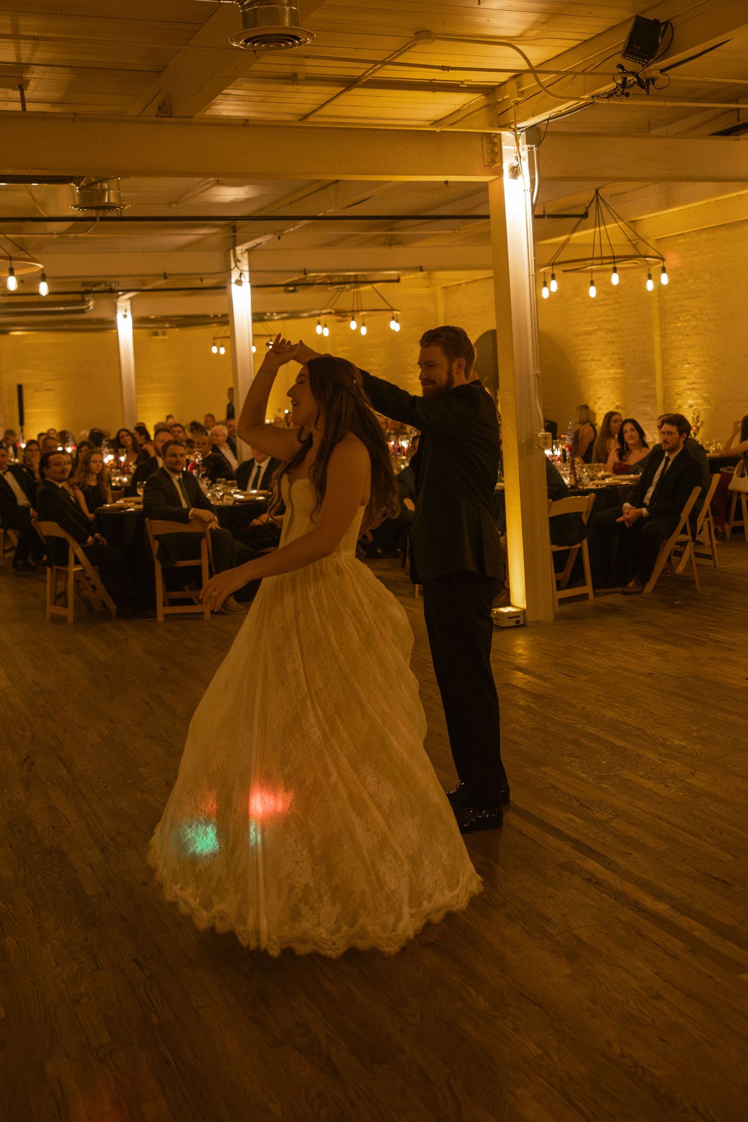 Wedding couple dancing in a warmly lit reception hall with guests seated in background.