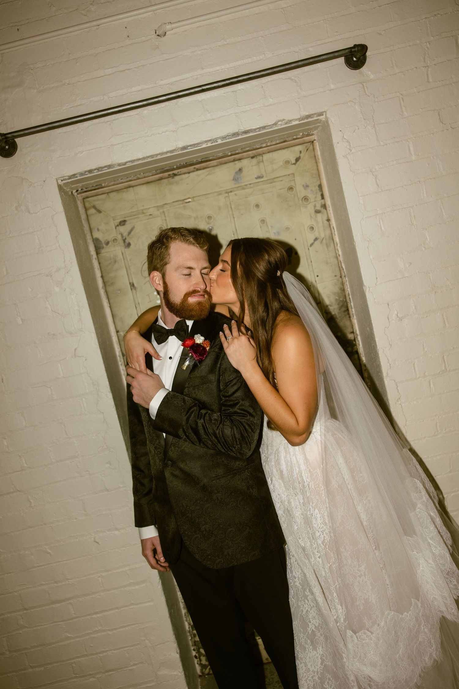A wedding couple shares a tender moment against a white brick wall in a vintage-style setting.