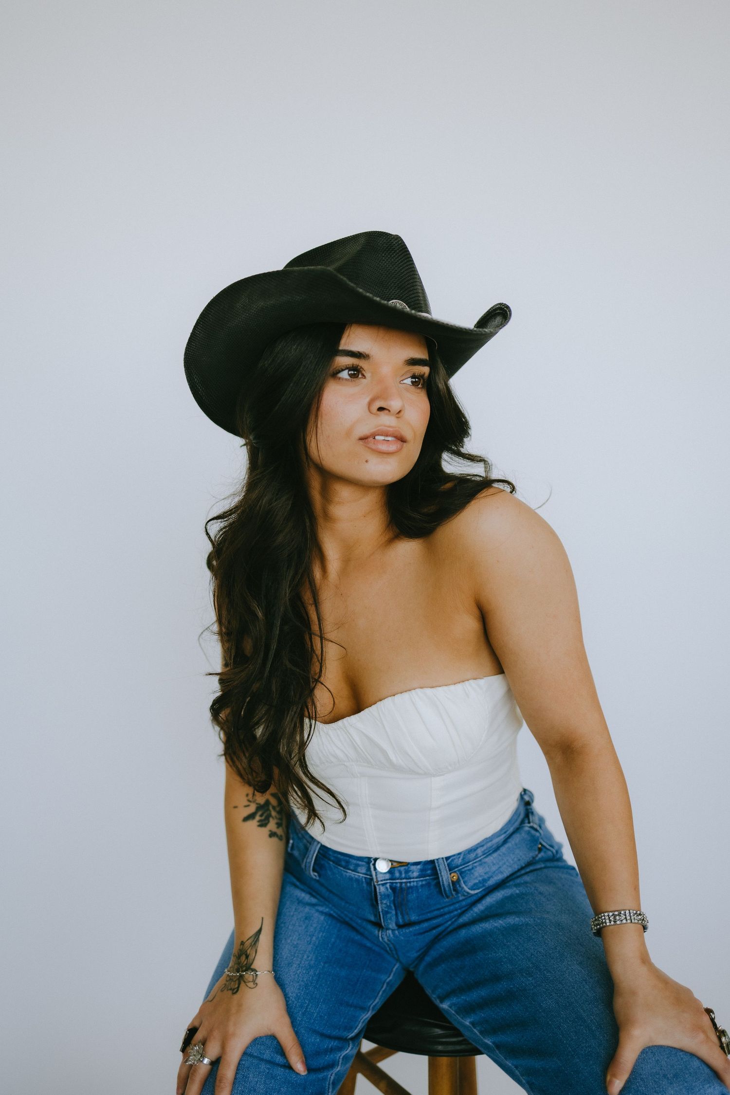 Fashion model poses on wooden stool wearing strapless top and denim against minimalist background.