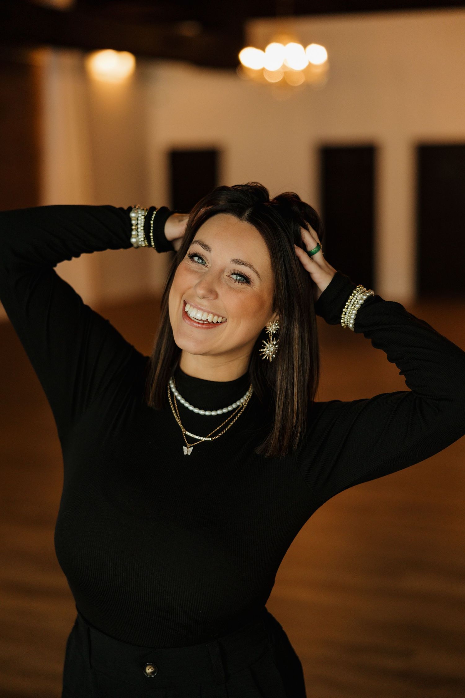 Fashionable portrait with sparkling jewelry and a black sweater in a warmly lit indoor setting.