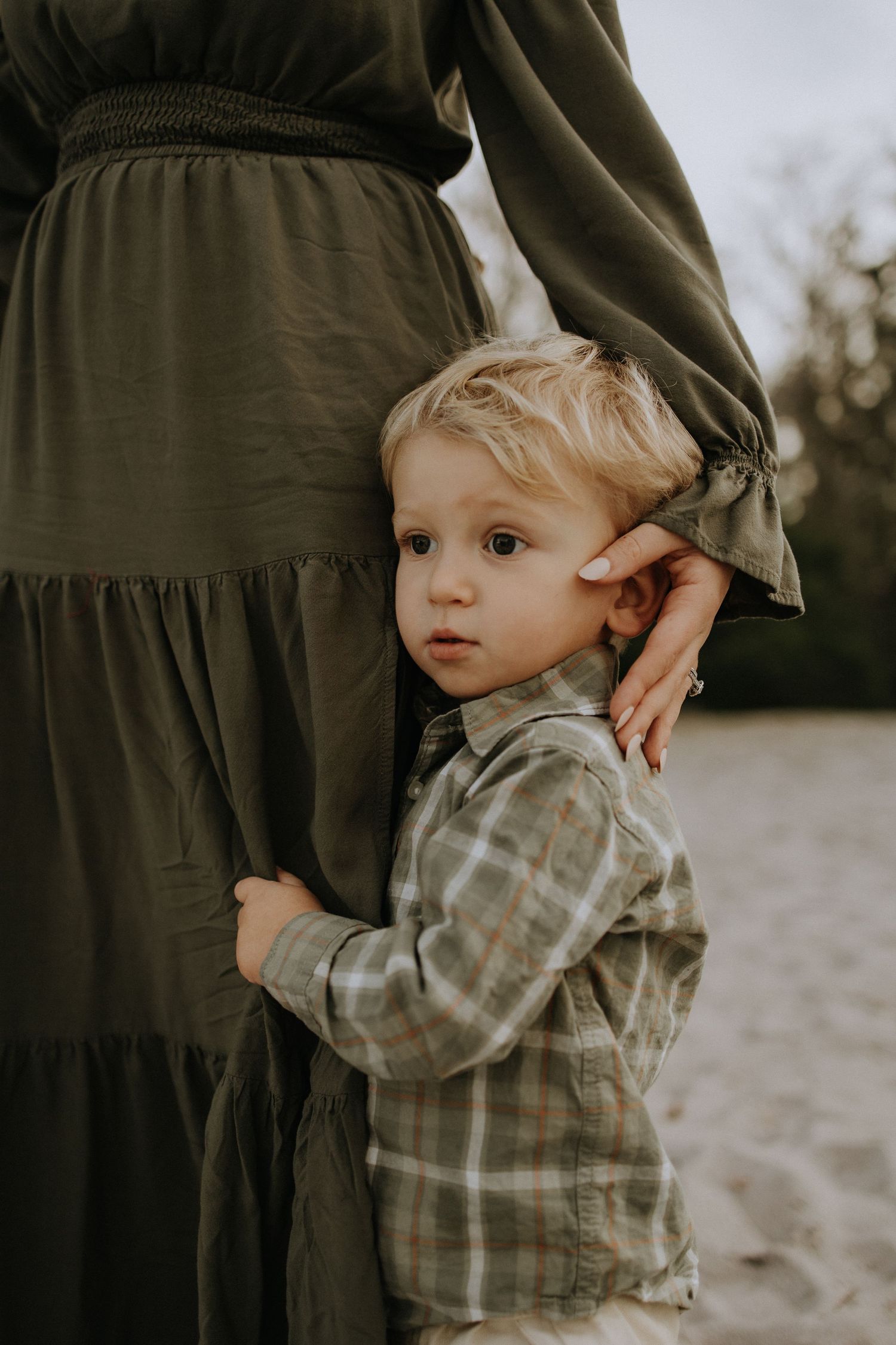 A small child in a plaid shirt cuddles close to someone wearing a dark dress on a beach.