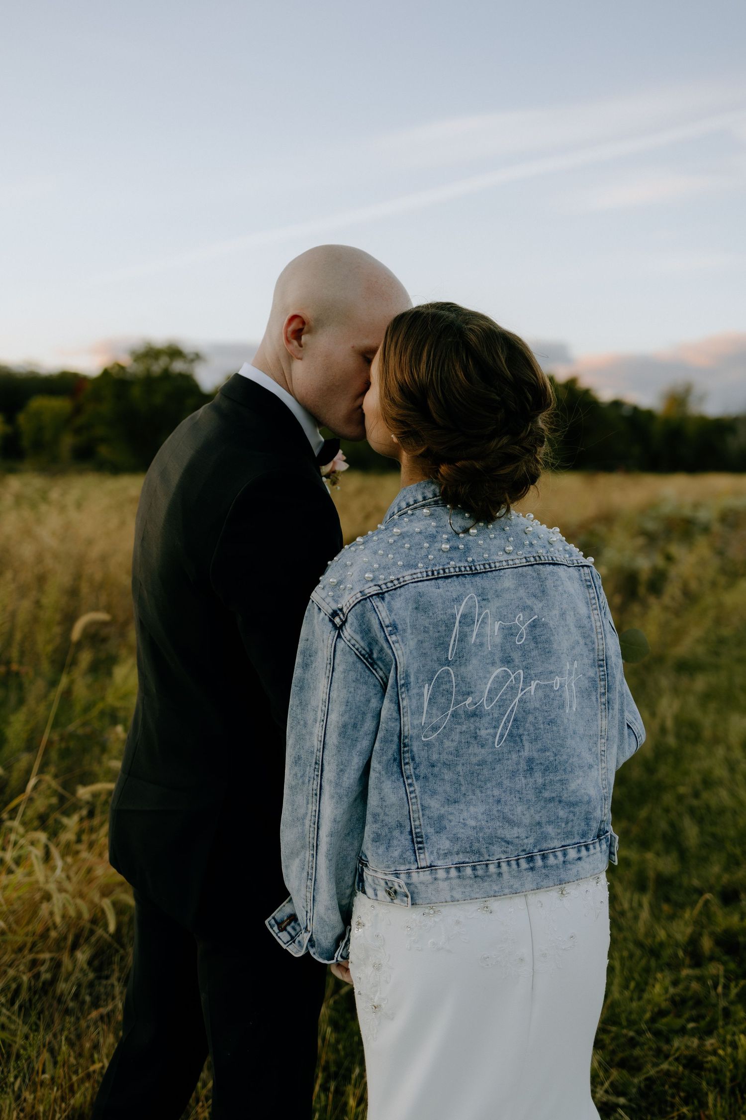 A couple shares a romantic moment in a field at sunset, with the bride wearing a customized denim jacket over her dress.