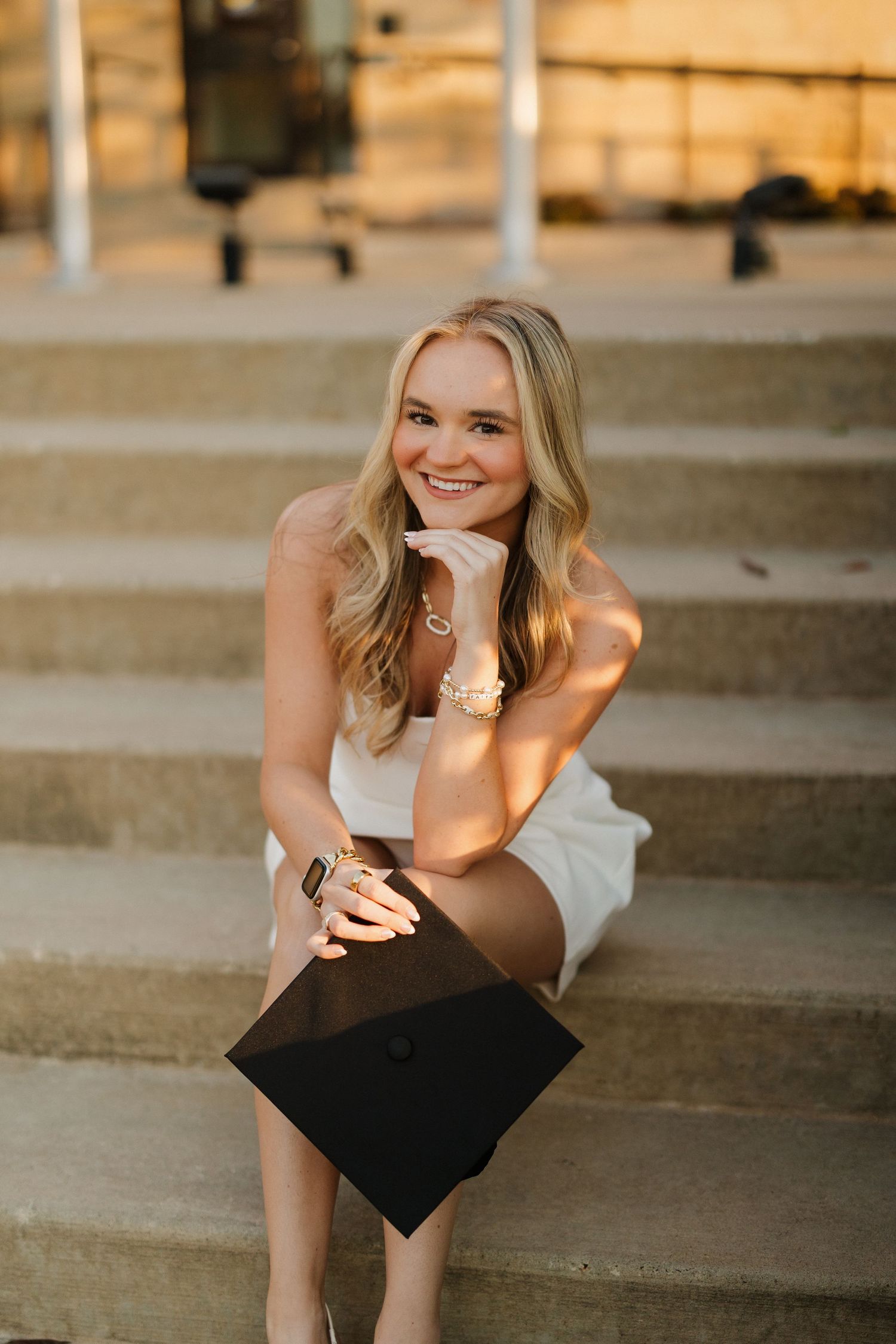Graduate in white dress sits on steps holding diploma during golden hour.