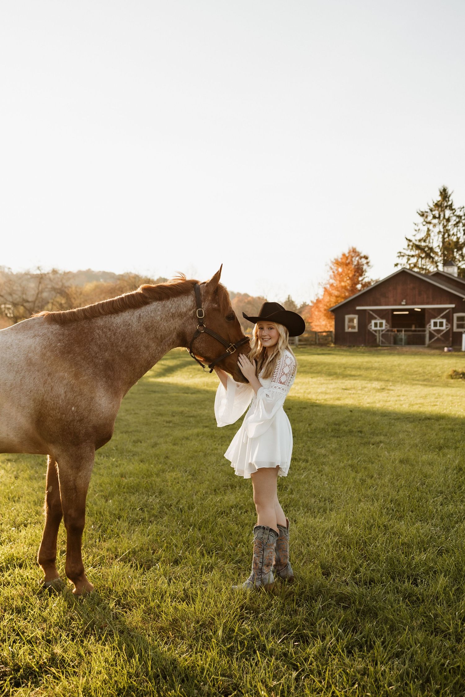 A person in a white dress pets a brown horse in a countryside setting during golden hour.