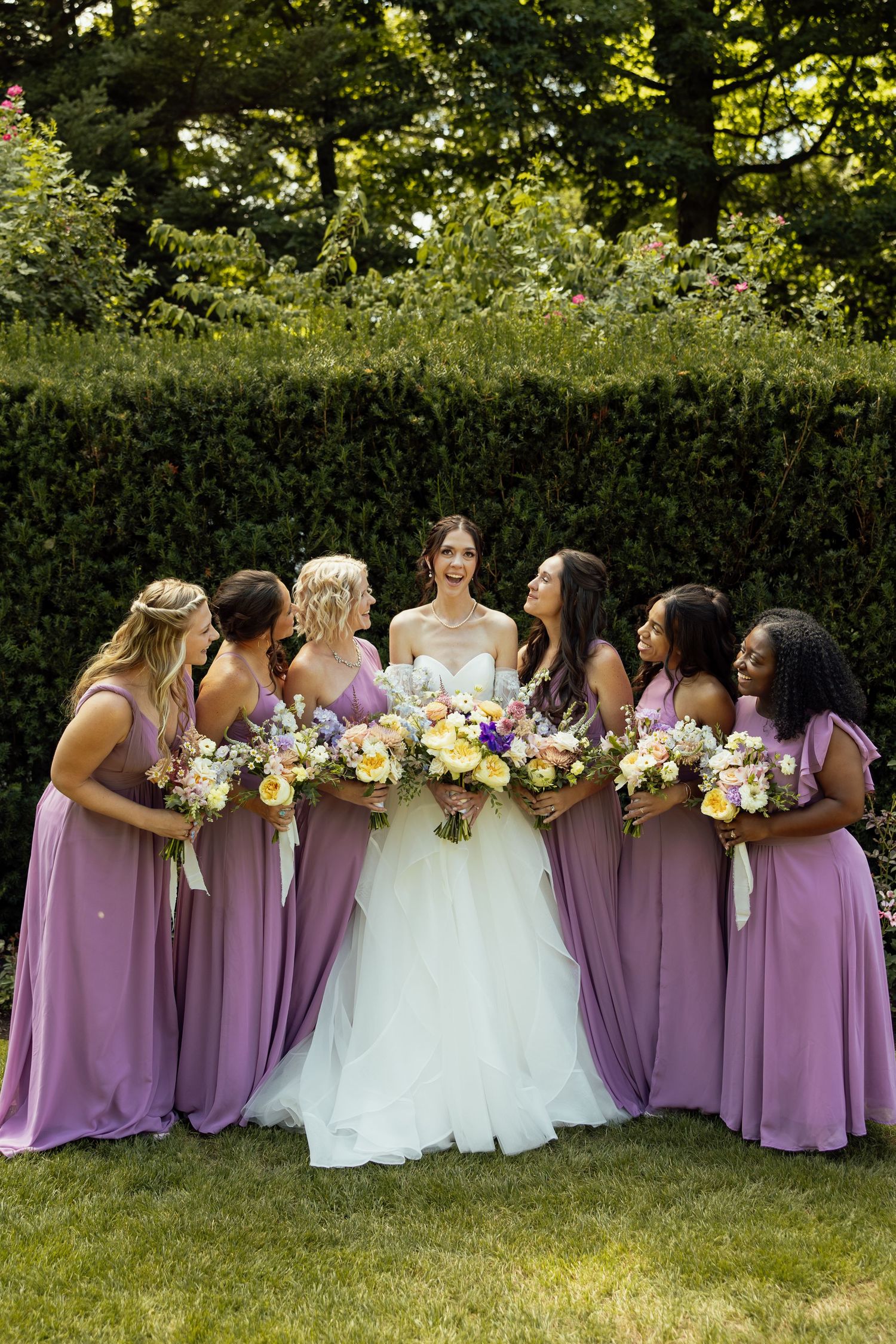 Wedding party in purple bridesmaid dresses holding colorful bouquets on a garden lawn.