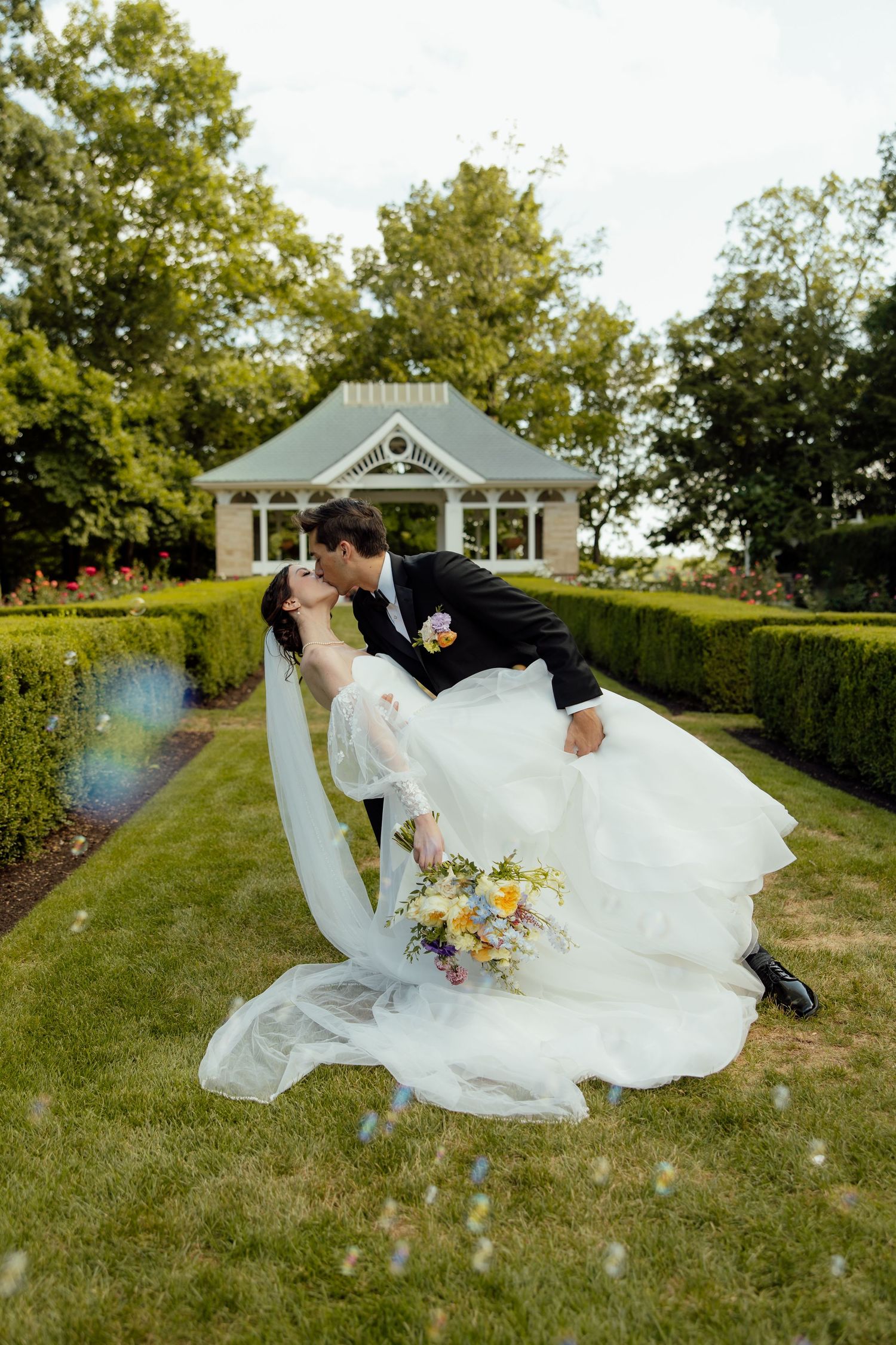 Romantic wedding couple embraces on grassy lawn with white flowing dress and veil creating elegant movement in sunlight.