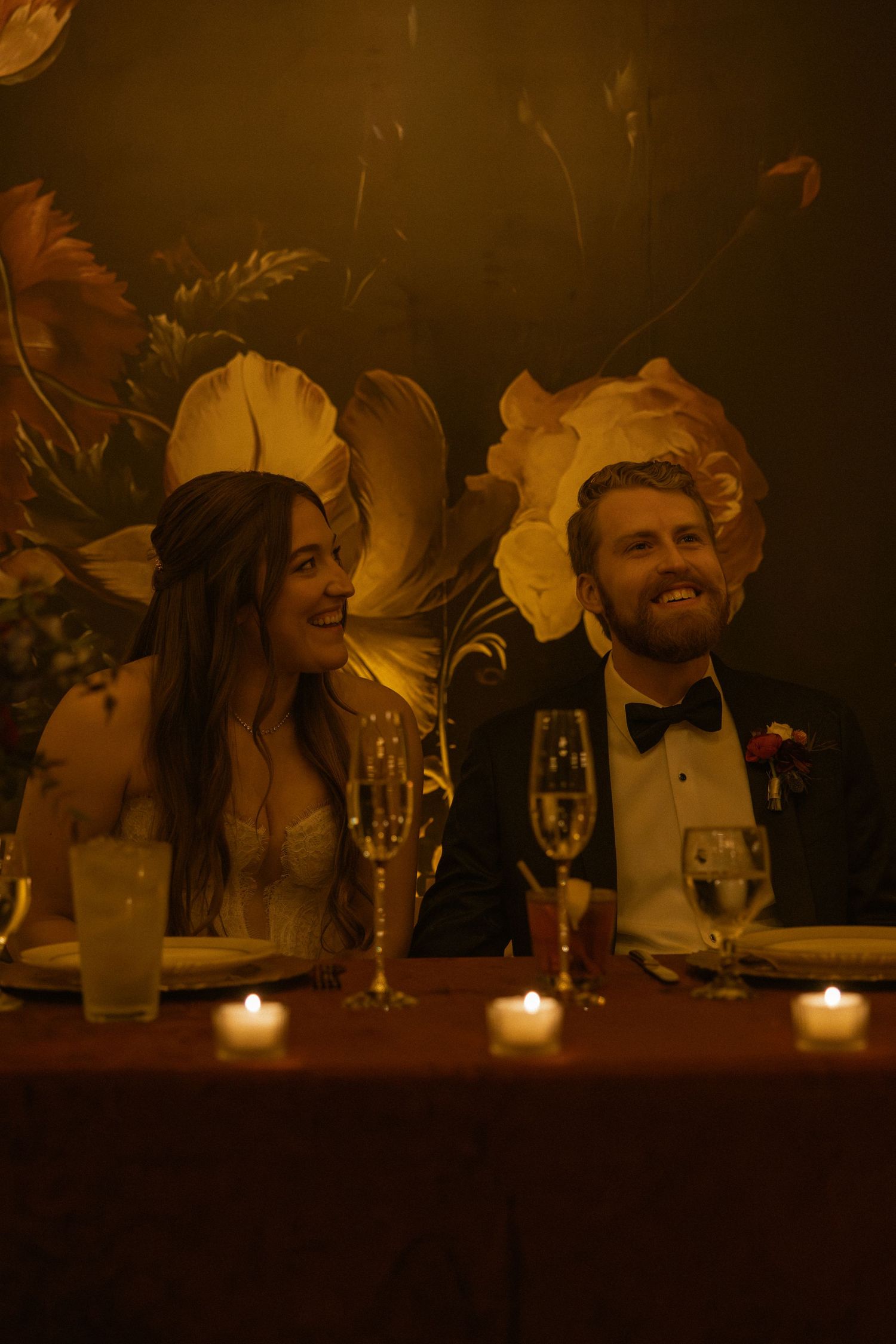 Wedding party guests laugh and smile at a candlelit reception table during an evening celebration.