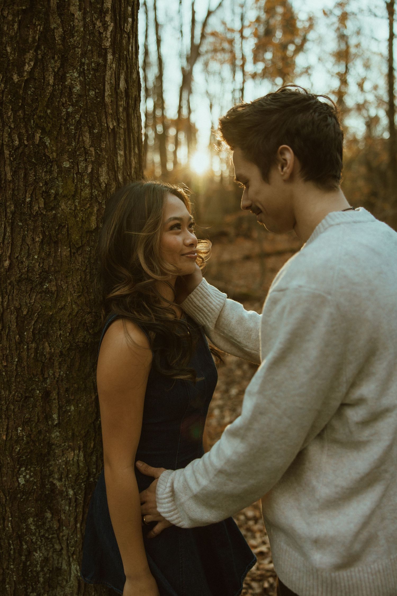 A couple shares an intimate moment together against a tree trunk during golden hour in an autumn forest setting.