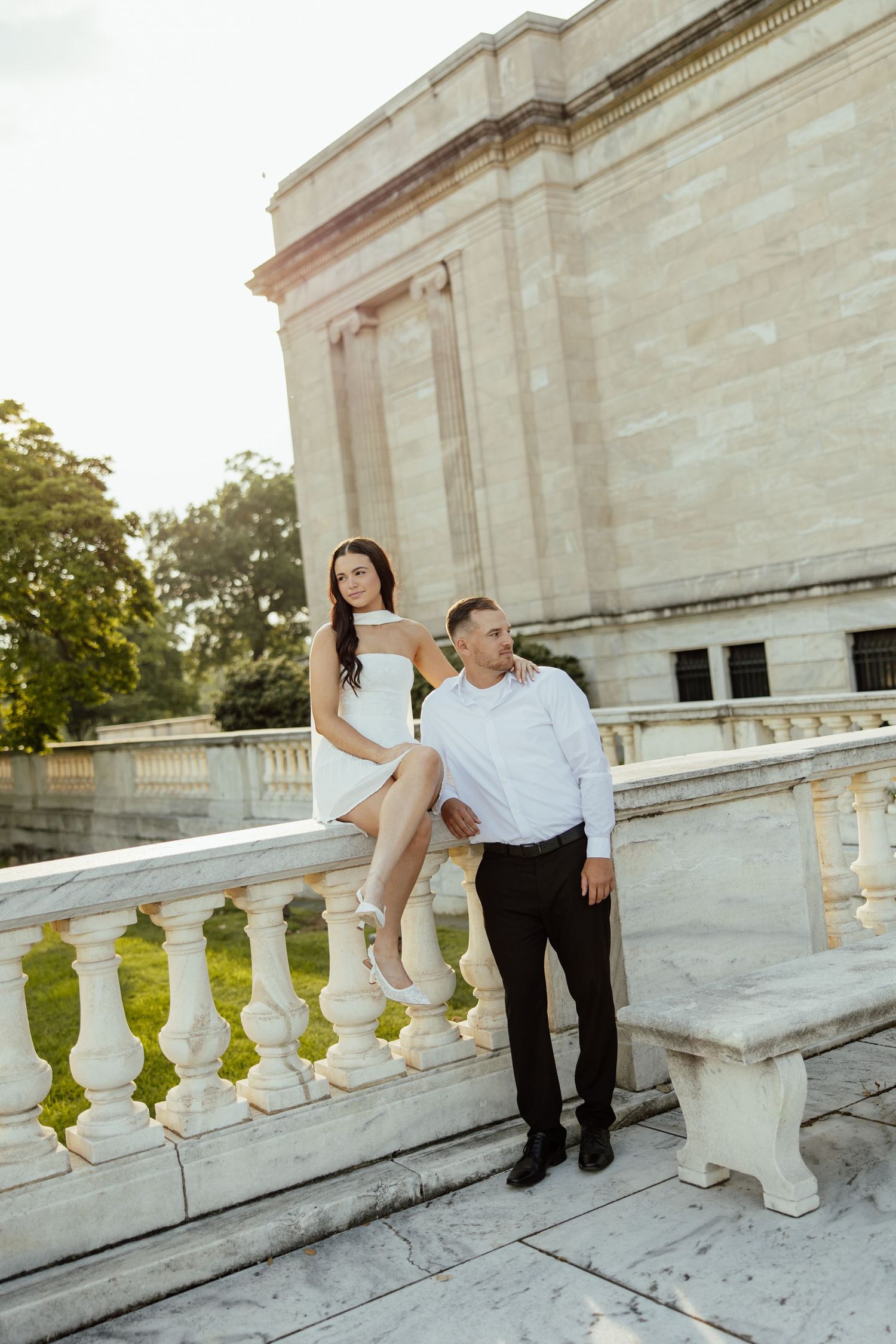 A couple in formal attire poses playfully on white stone steps of a historic building during a photoshoot.