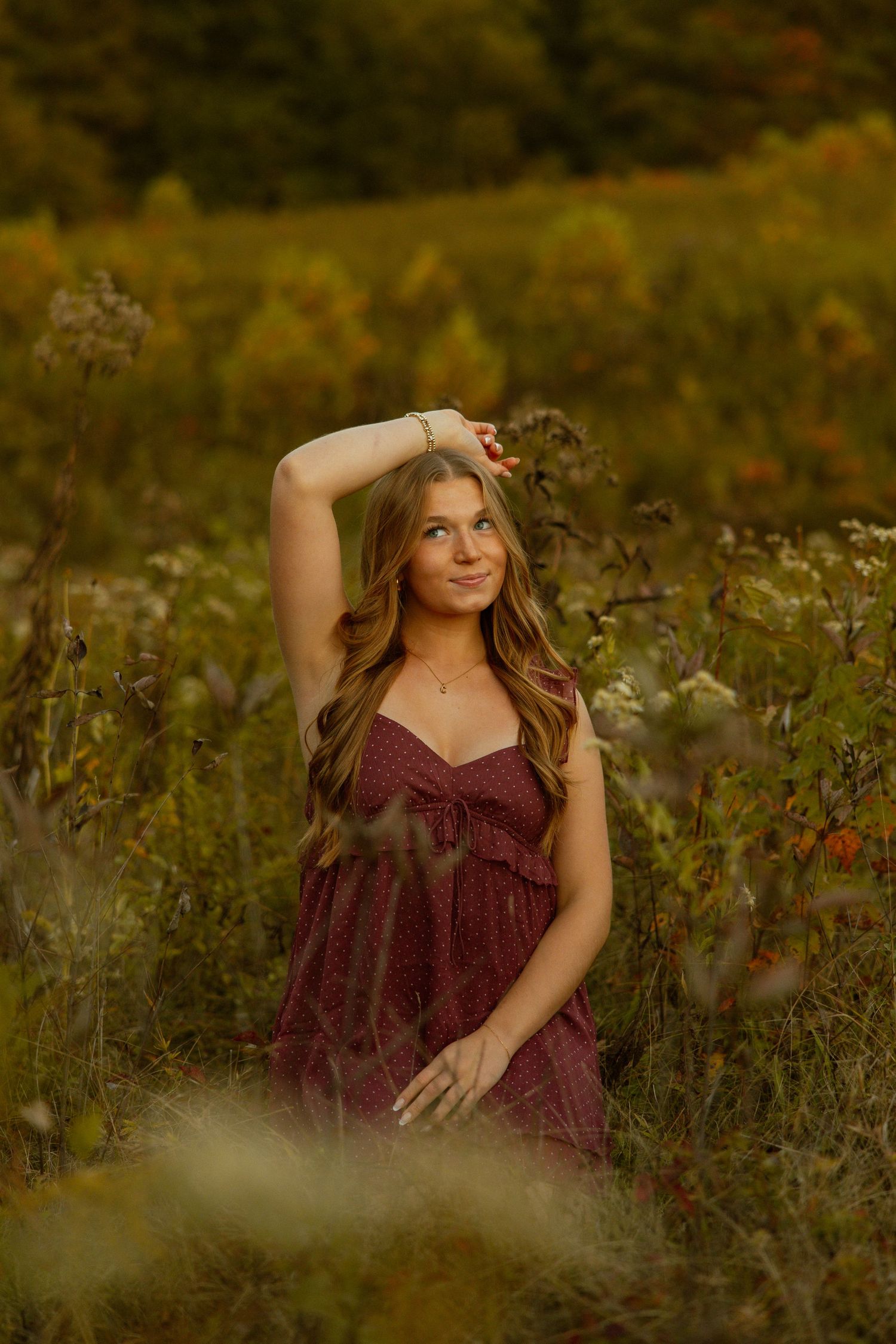 A person in a burgundy dress poses gracefully among wildflowers in a golden autumn meadow during sunset.
