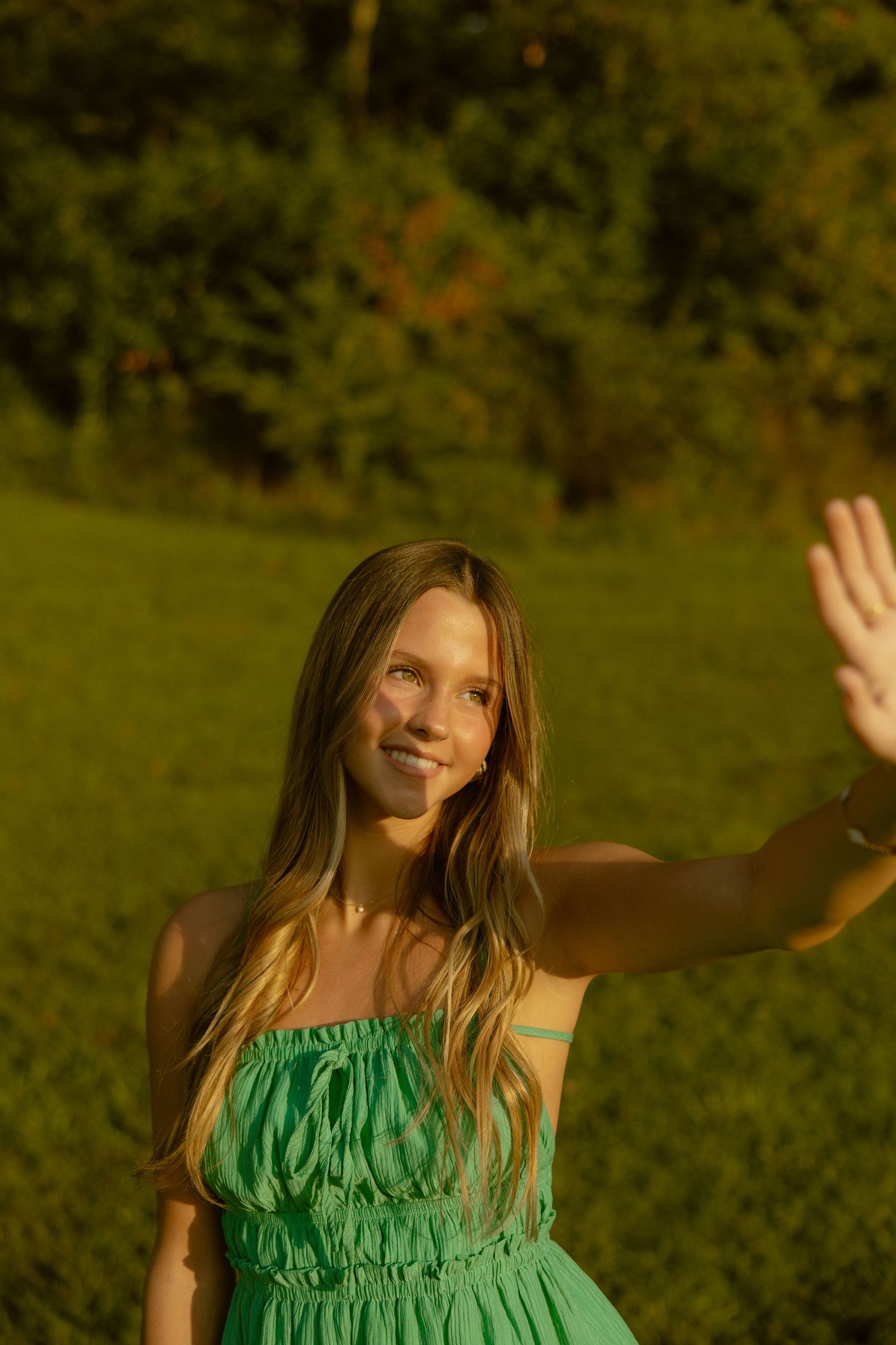 Person in green ruffled dress dances joyfully in a sunlit outdoor setting with natural greenery in background.