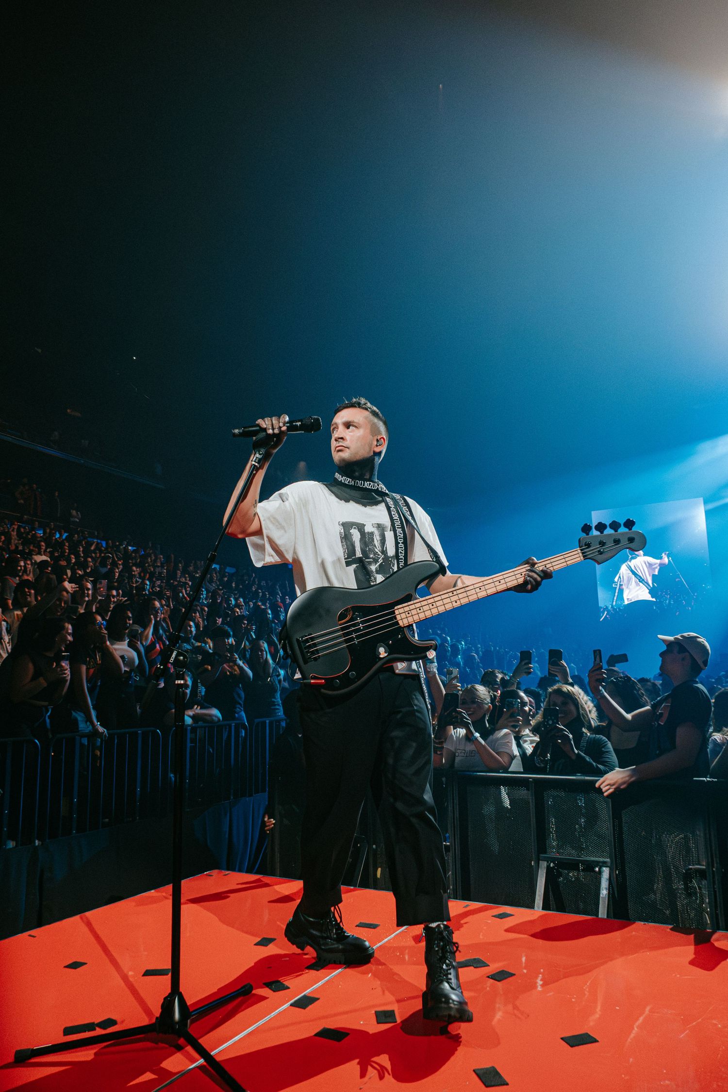 Performer with bass guitar stands on red stage under blue lighting during concert.
