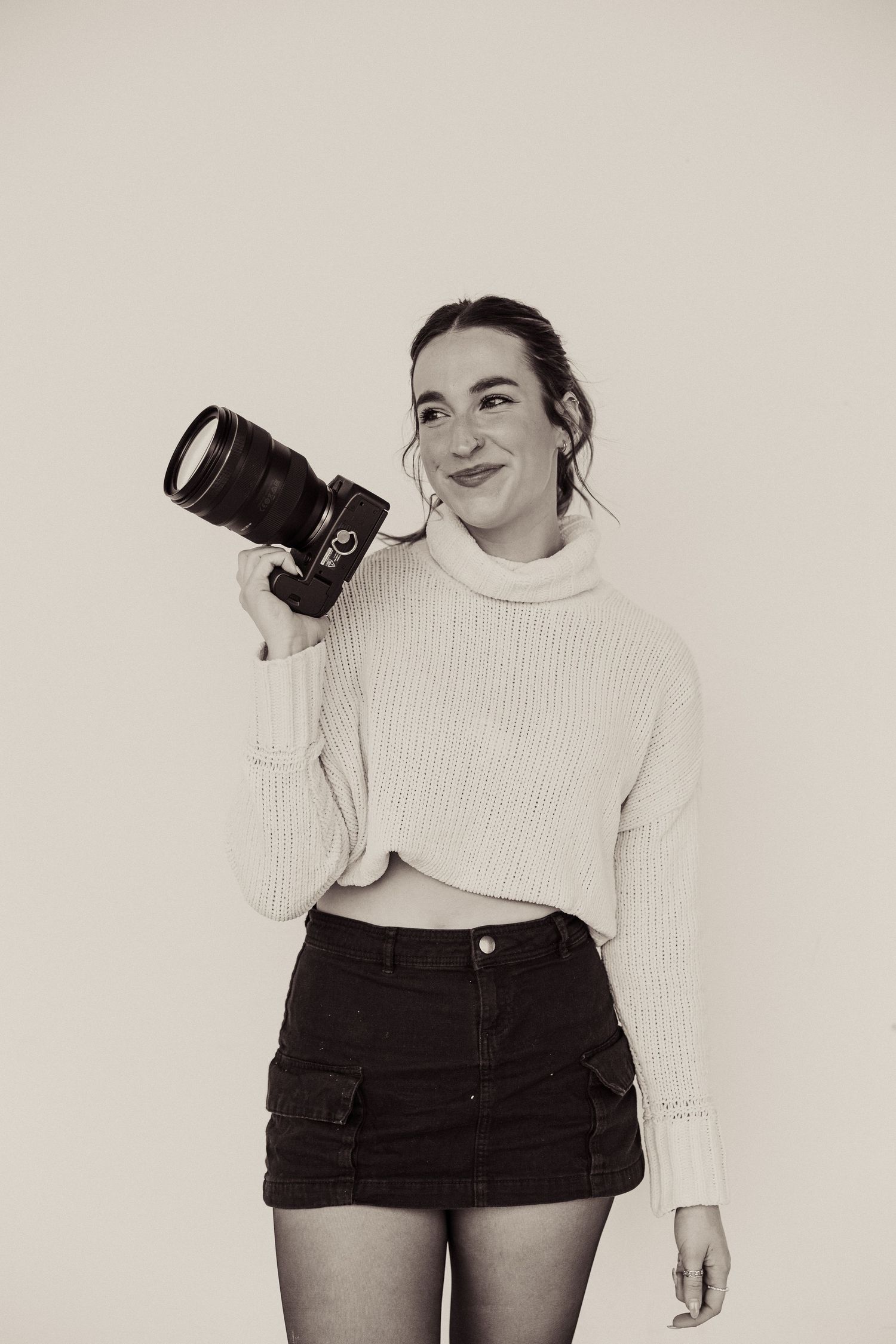 A photographer in a cream sweater and dark shorts holds a professional camera in a studio setting.