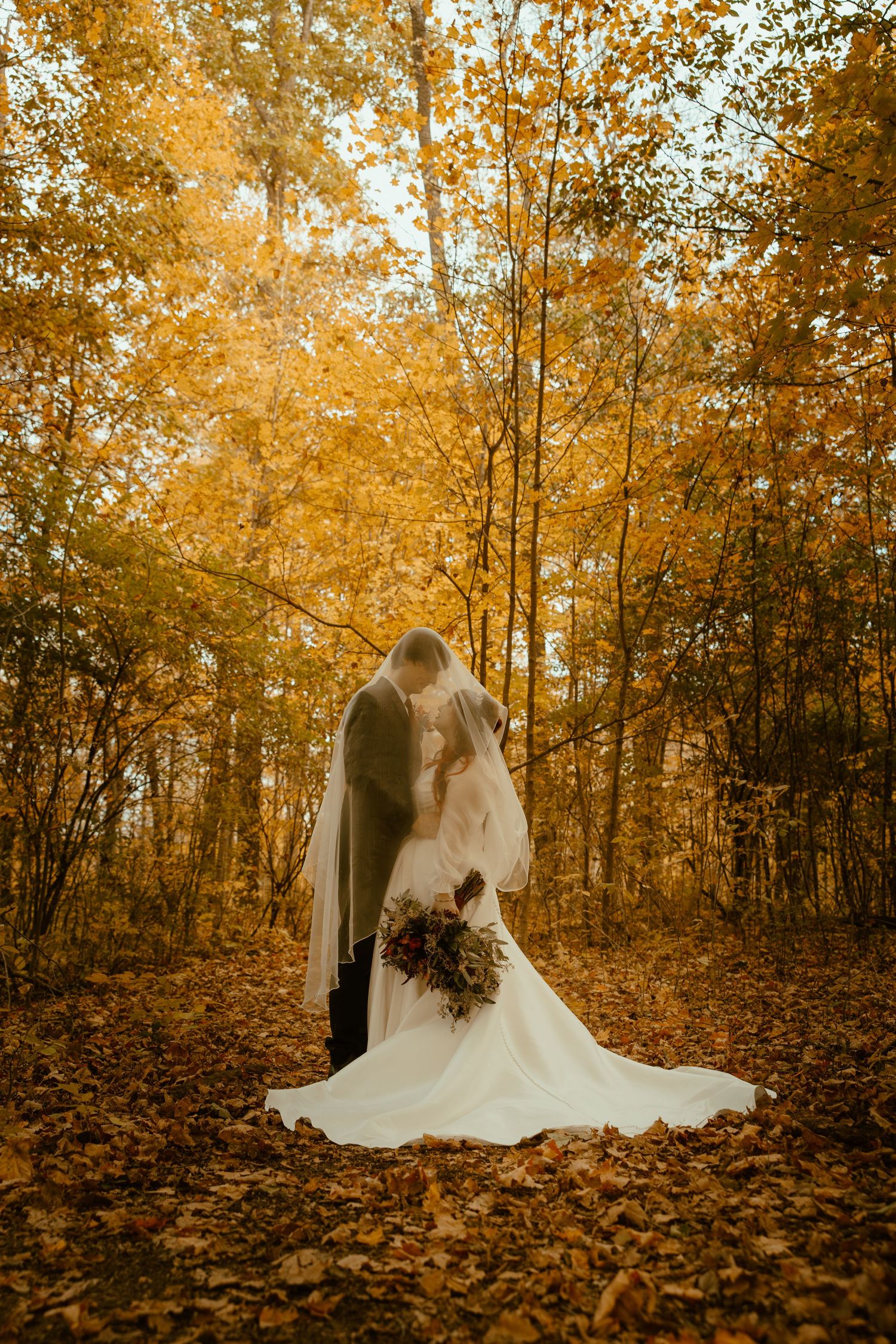 A romantic autumn wedding scene in a forest with golden leaves and a flowing white dress against warm fall foliage.