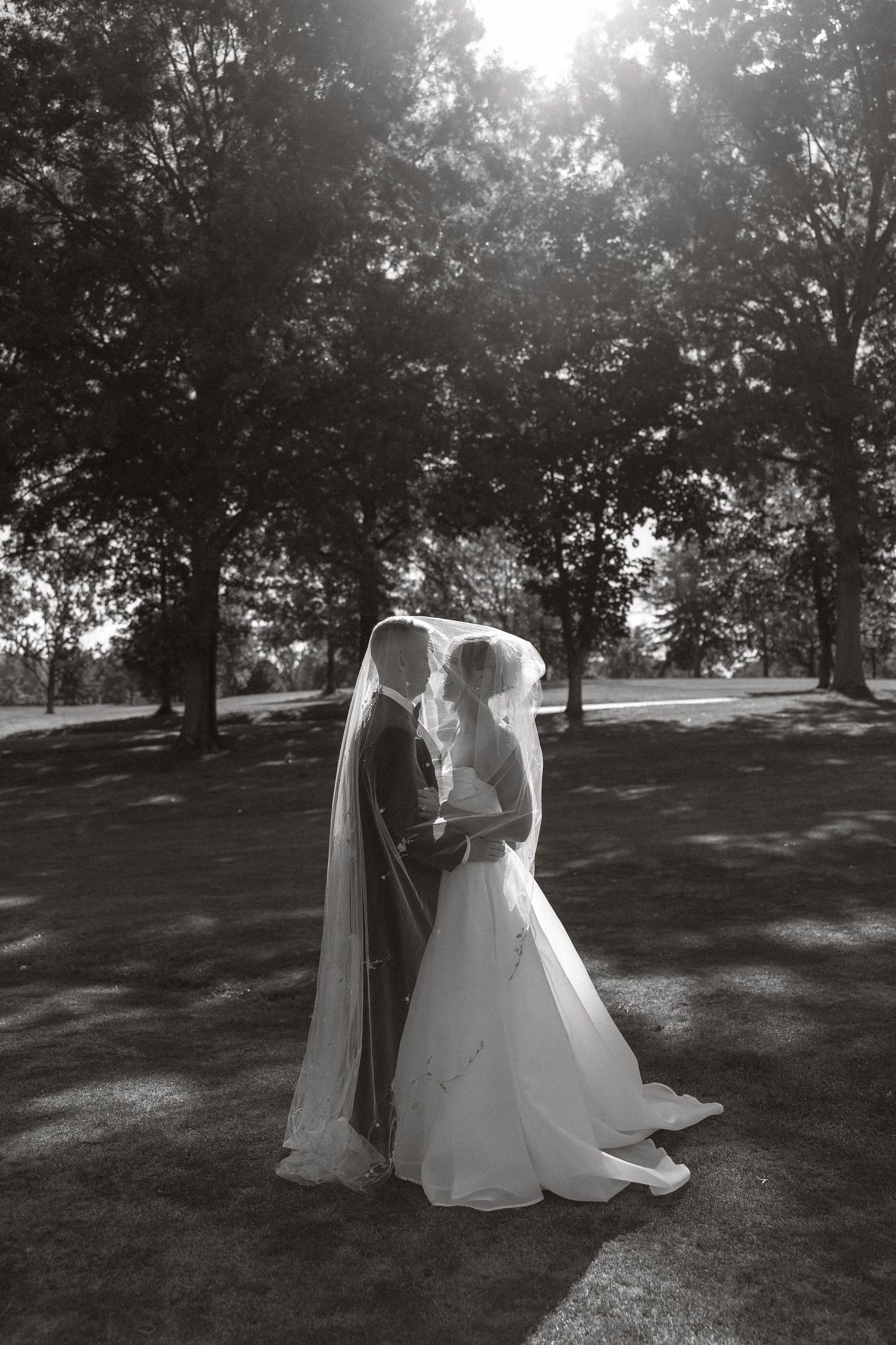 Black and white photo of a wedding couple dancing in a sunlit park with trees casting shadows.