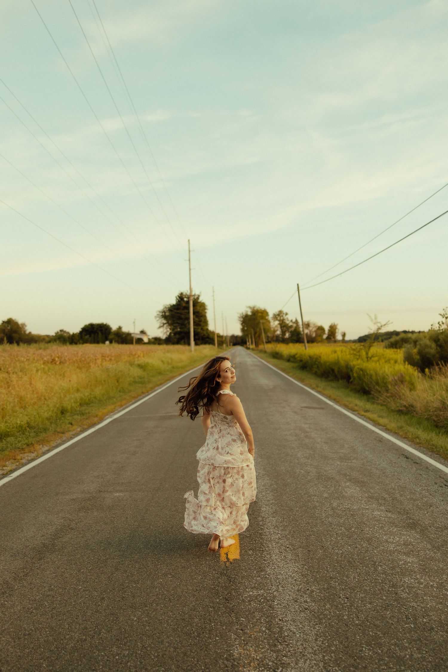 A person in a floral dress walks down a rural road surrounded by fields on a sunny day.