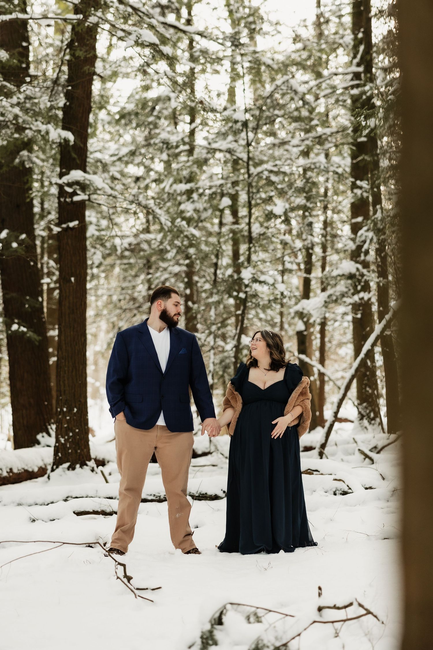 A couple shares a candid moment while standing in a snow-covered forest wearing formal attire.