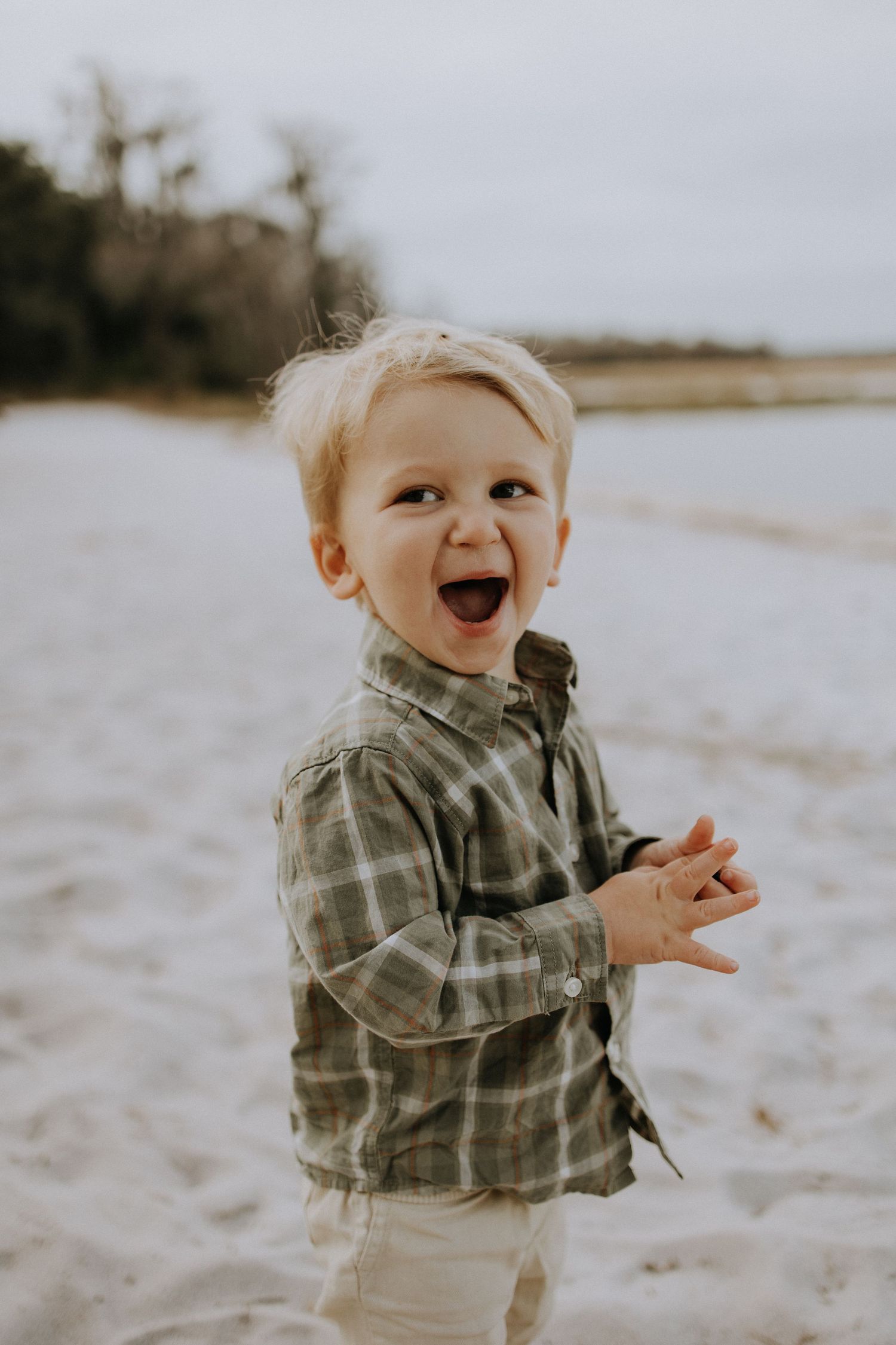 A small child wearing a plaid shirt laughs with delight on a beach.