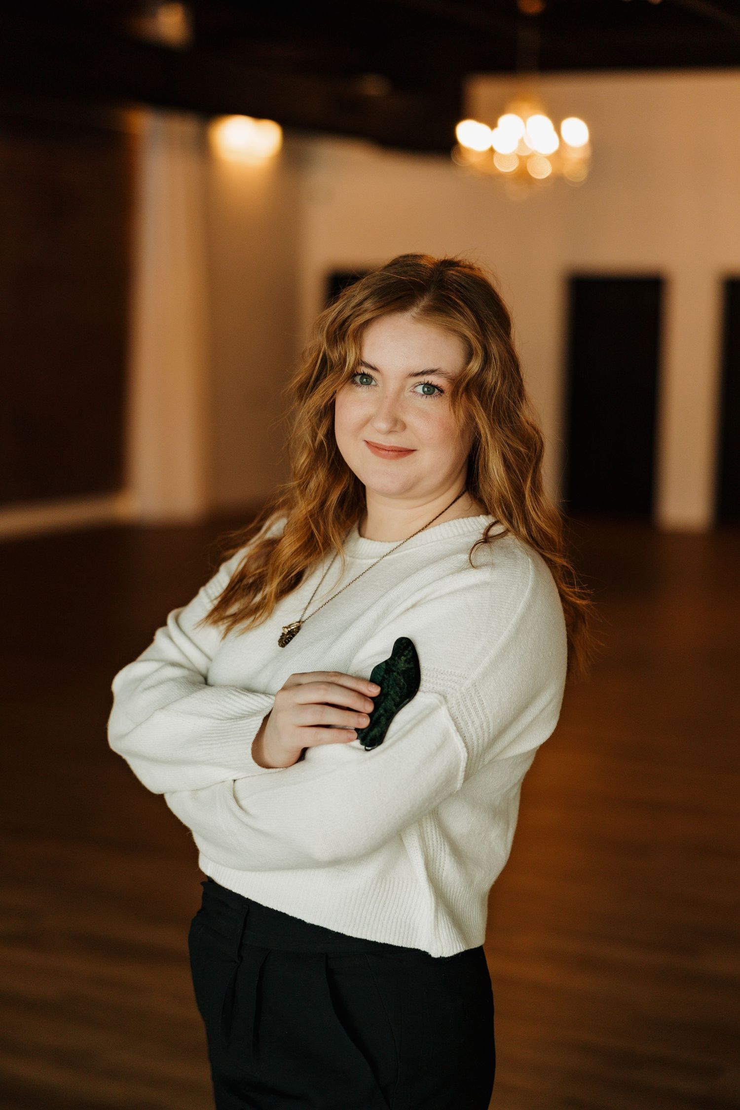 Professional portrait in a white sweater standing confidently against a warm interior backdrop with bokeh lighting.