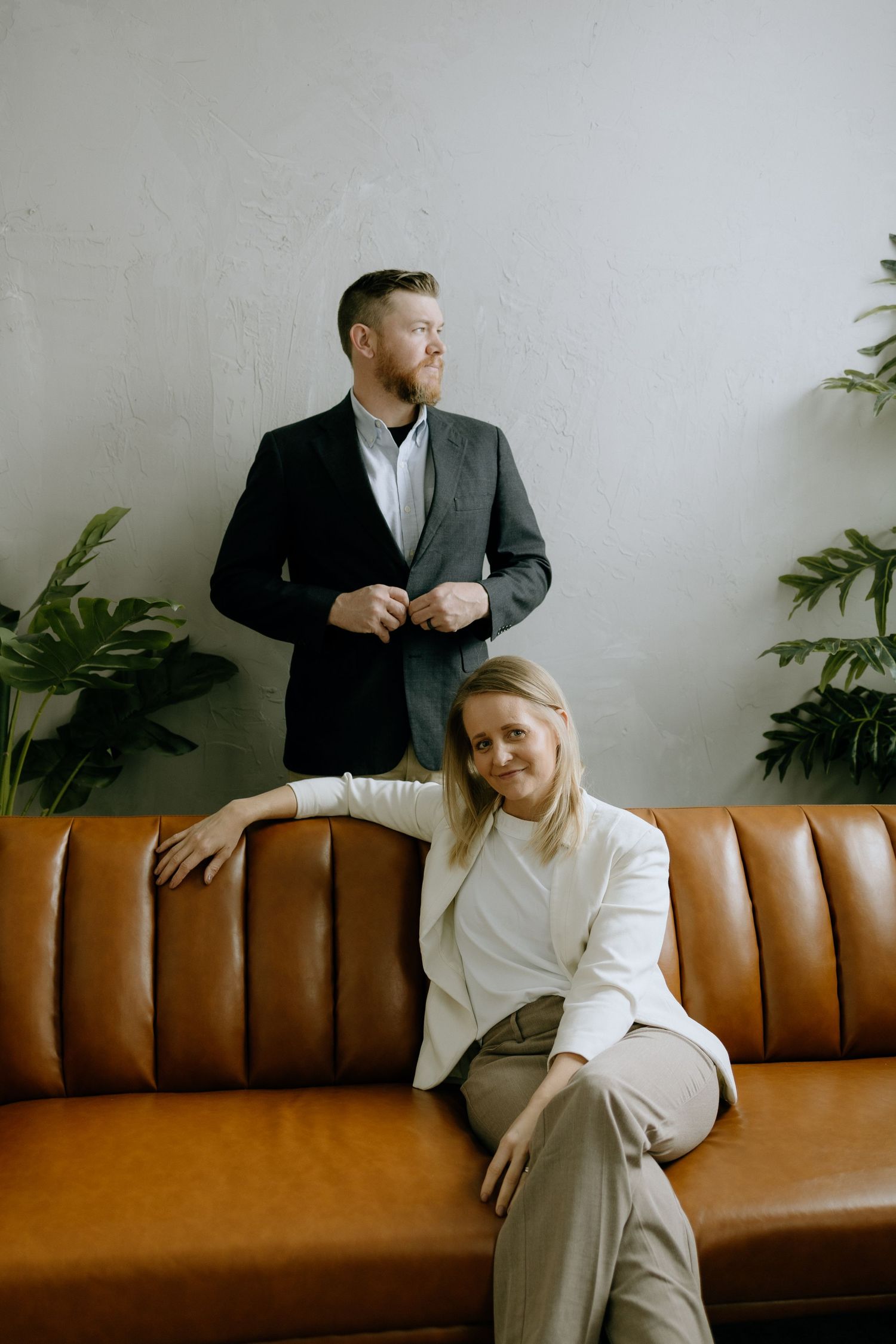 Two professionals posing on brown leather couch with greenery and minimalist background.