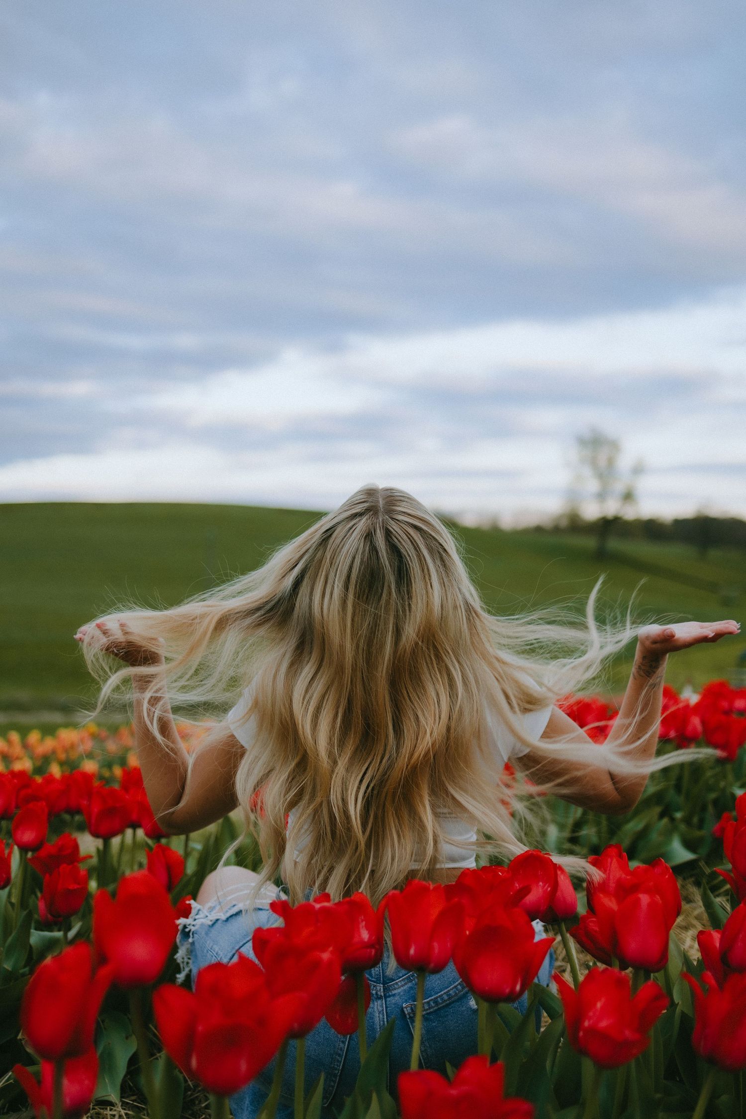 Blonde hair blowing in wind among red tulips under cloudy sky.