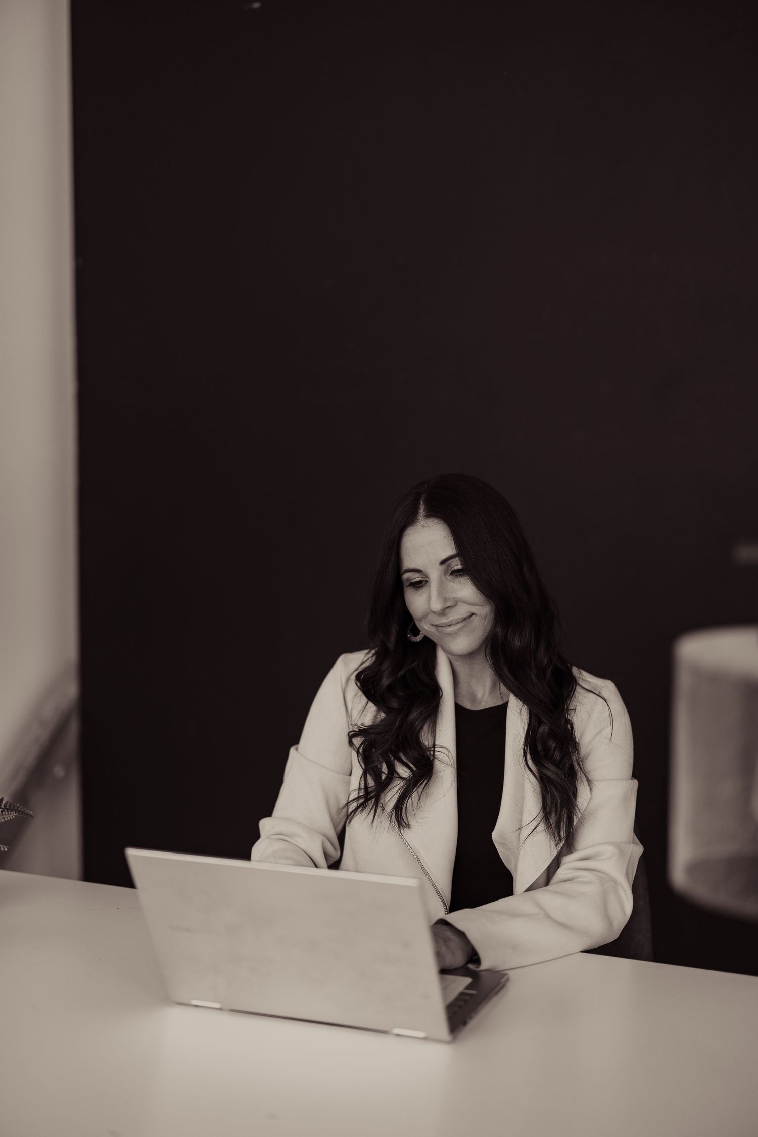 Professional at desk working on laptop wearing white blazer in black and white photo.
