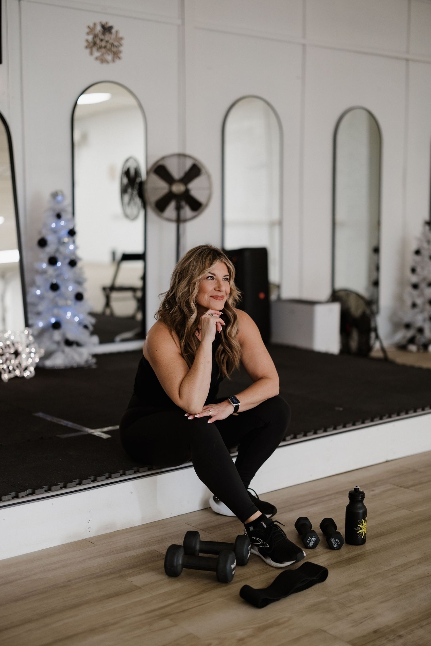 Woman in black workout attire sits thoughtfully near exercise equipment in a mirrored studio space.
