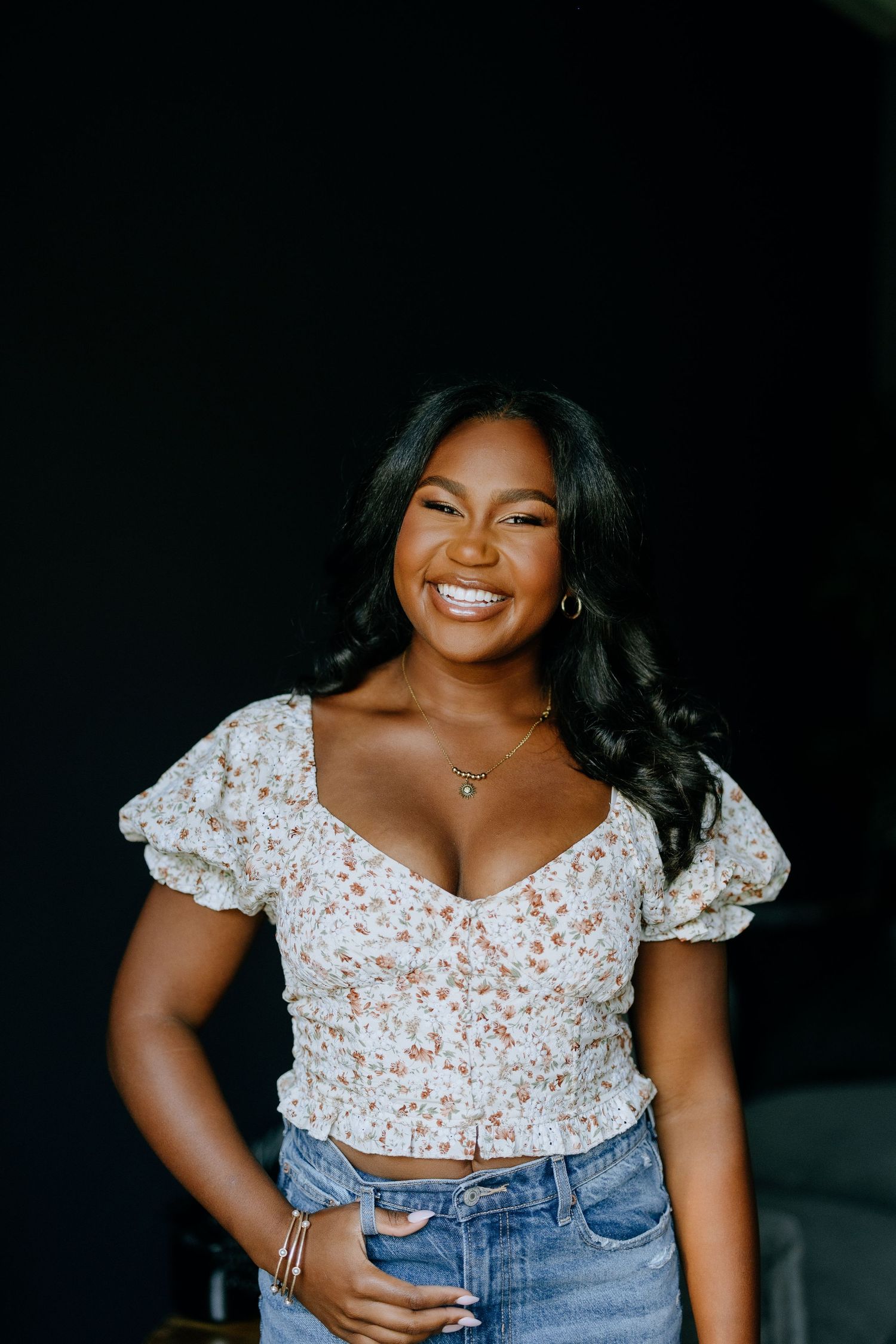 Studio portrait wearing floral crop top and denim against dark background.