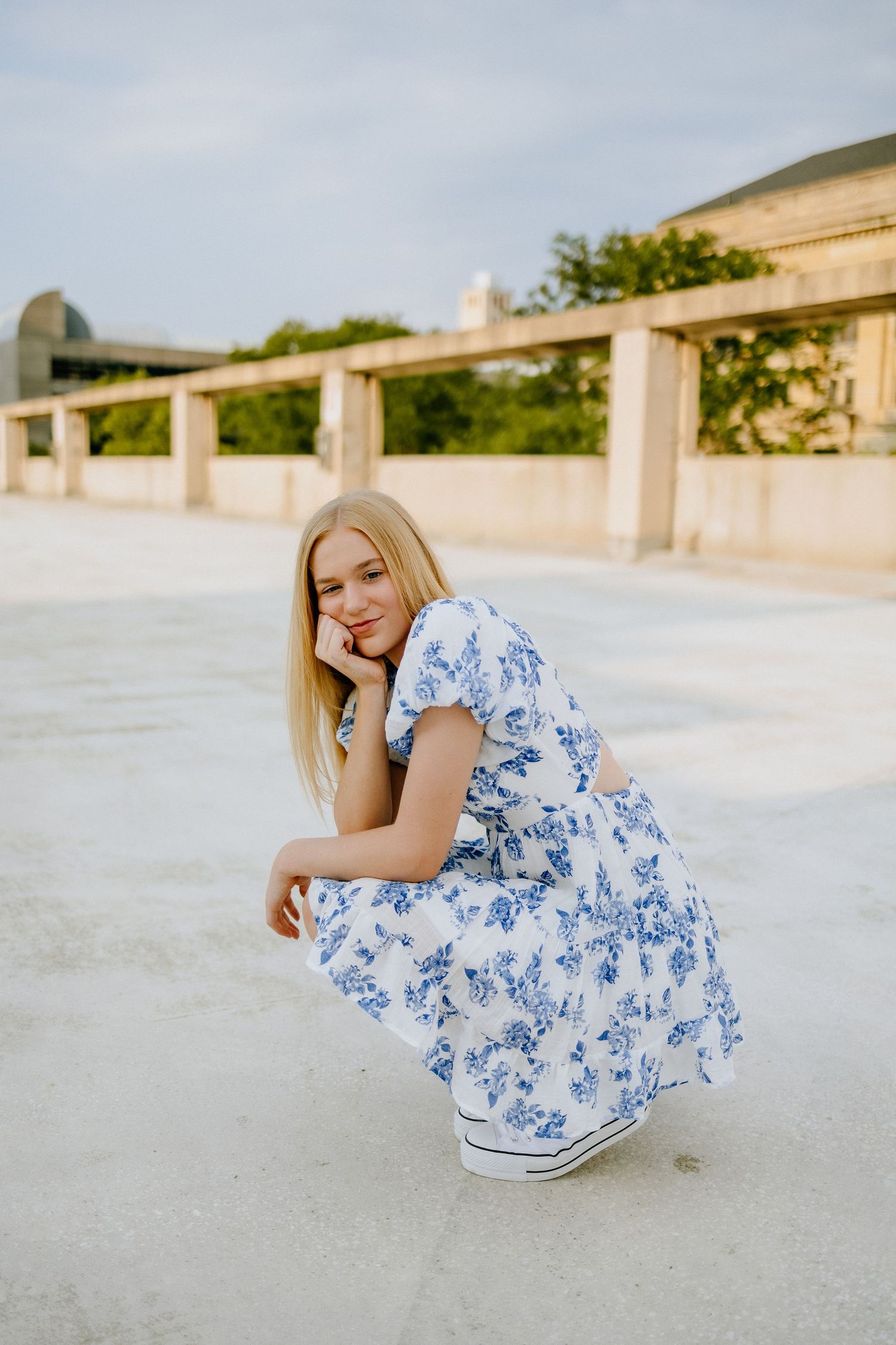 Woman in blue floral dress crouches while wearing white sneakers against concrete backdrop.